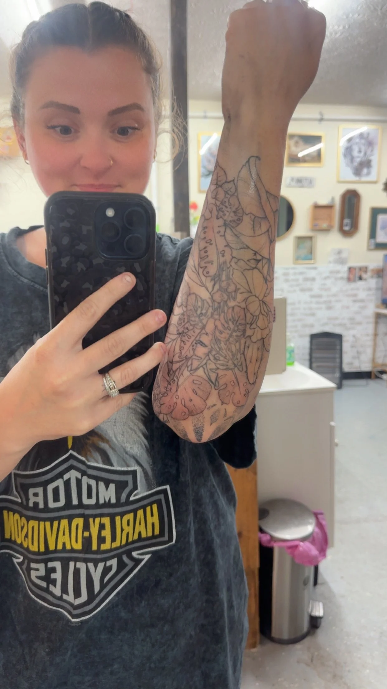 A woman taking a selfie in a mirror, showing her arm with a tattoo design in progress. She is smiling and has light brown hair pulled back, wearing a dark Harley-Davidson t-shirt.