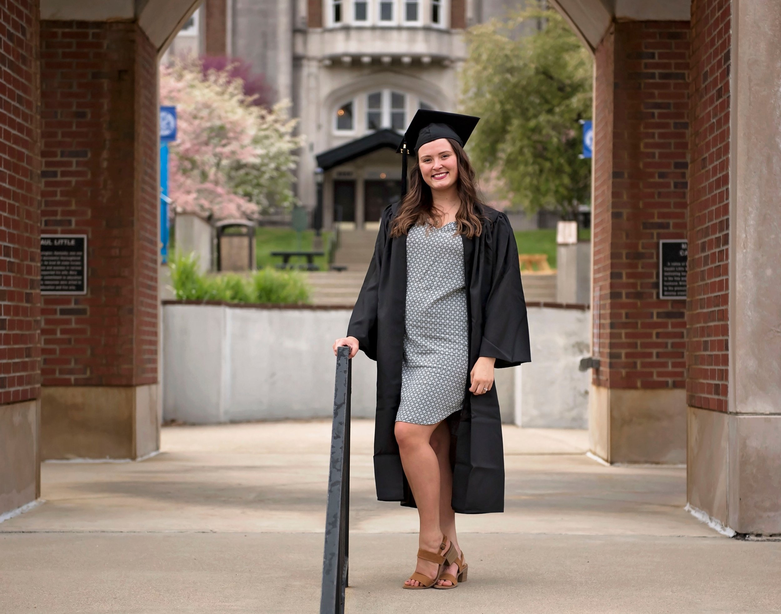 A young woman in a graduation cap and gown standing outdoors on a university campus, smiling and holding onto a railing with one hand.