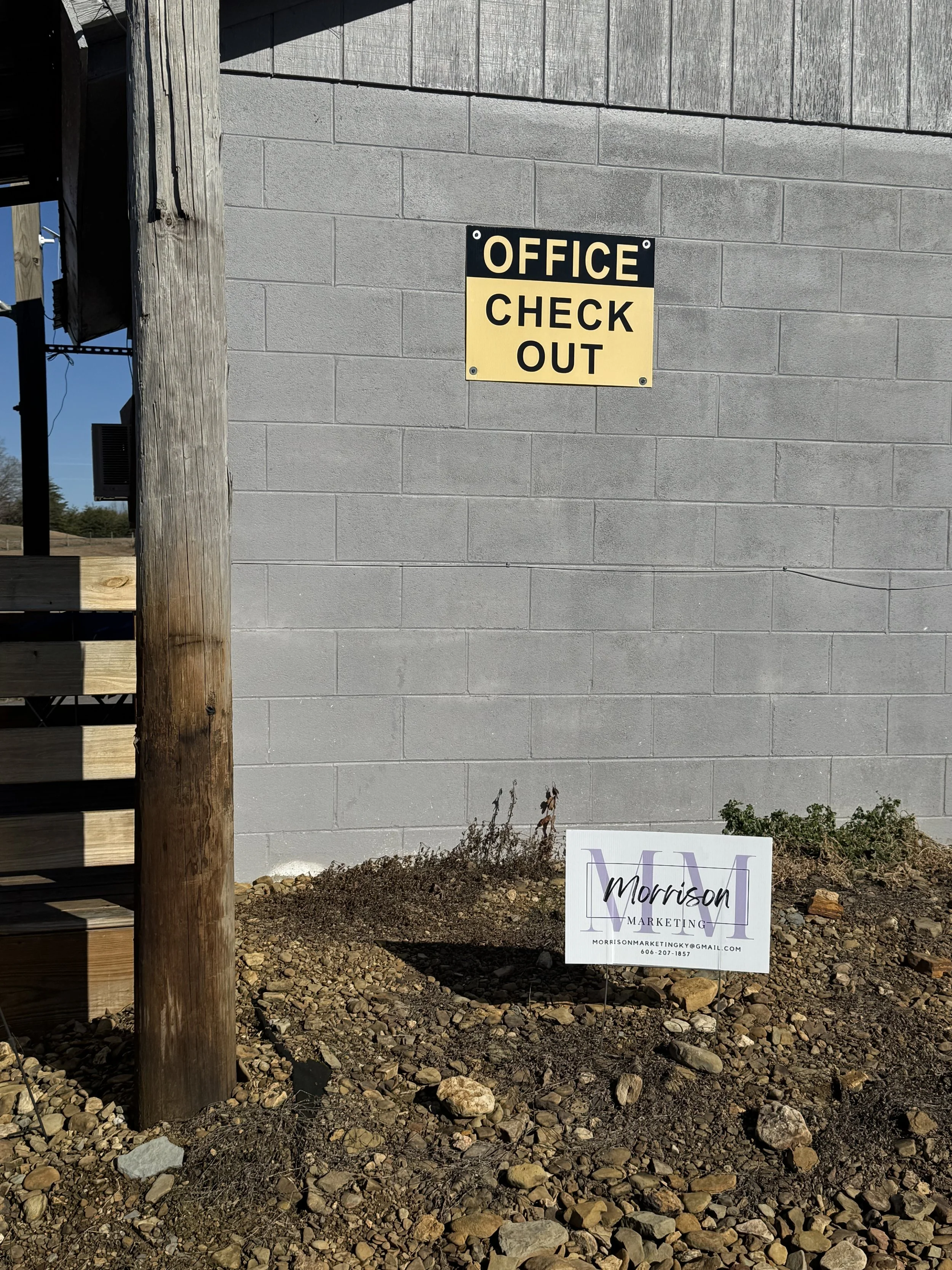 Sign on a gray brick wall that reads 'Office Check Out'. Small sign in dirt below the wall with 'M Morrison Marketing' and contact details.