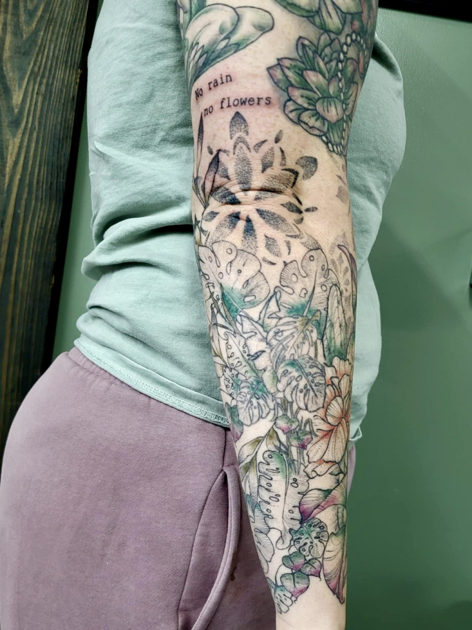 Close-up of a person's arm with a colorful floral tattoo and a quote reading 'No rain no flowers'.