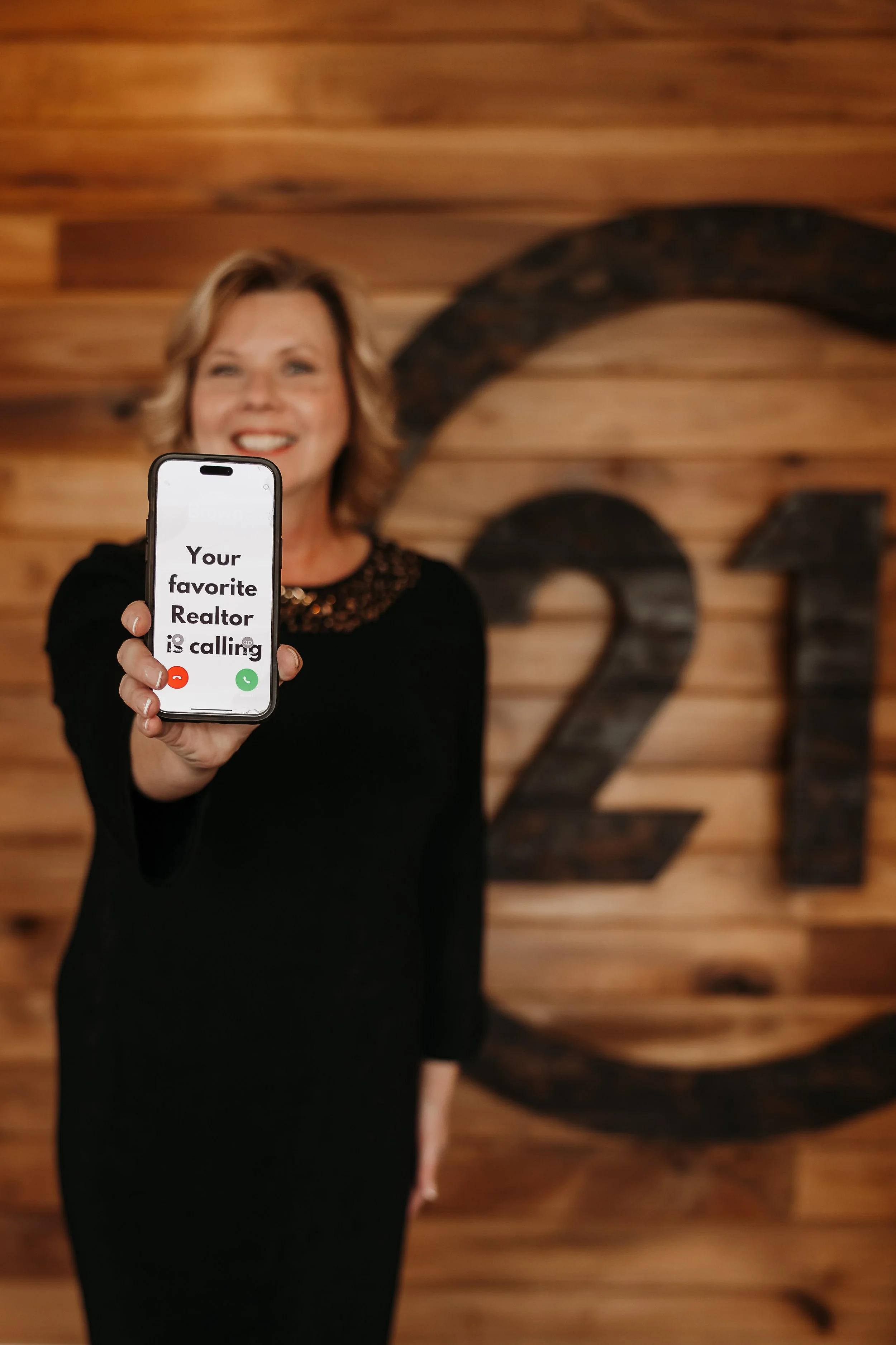 A woman smiling and holding a phone with an incoming call screen that reads "Your favorite Realtor is calling" in front of a wooden wall with large black numbers '21'.