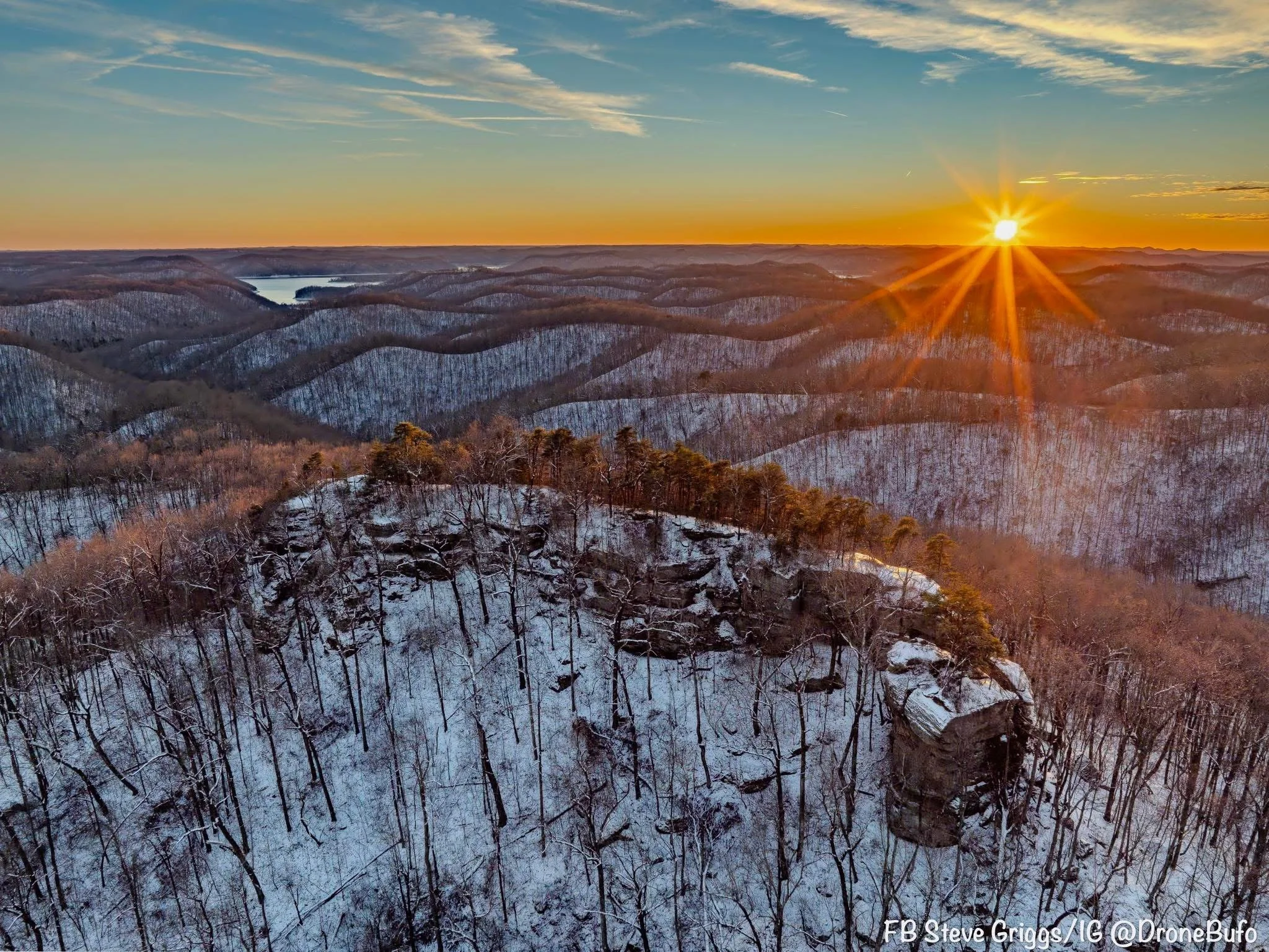 A winter landscape showing snow-covered forested hills and a rocky cliff with trees, with the sun setting on the horizon, casting a warm orange glow.