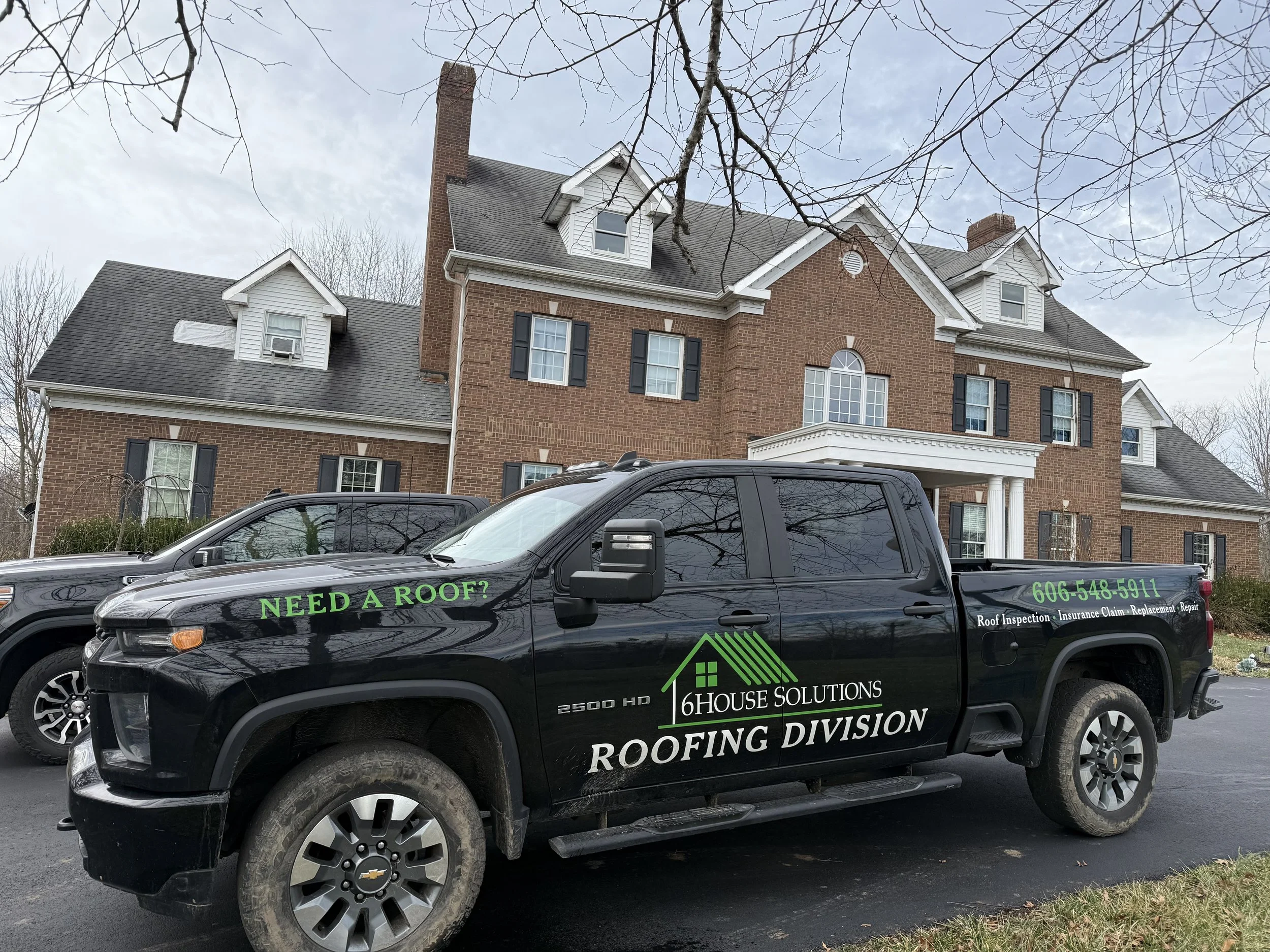 A black pickup truck parked in front of a large brick house. The truck has green and white lettering advertising a roofing company called 6 House Solutions, including about roof inspection, insurance claim, replacement, and repair services, with a ph