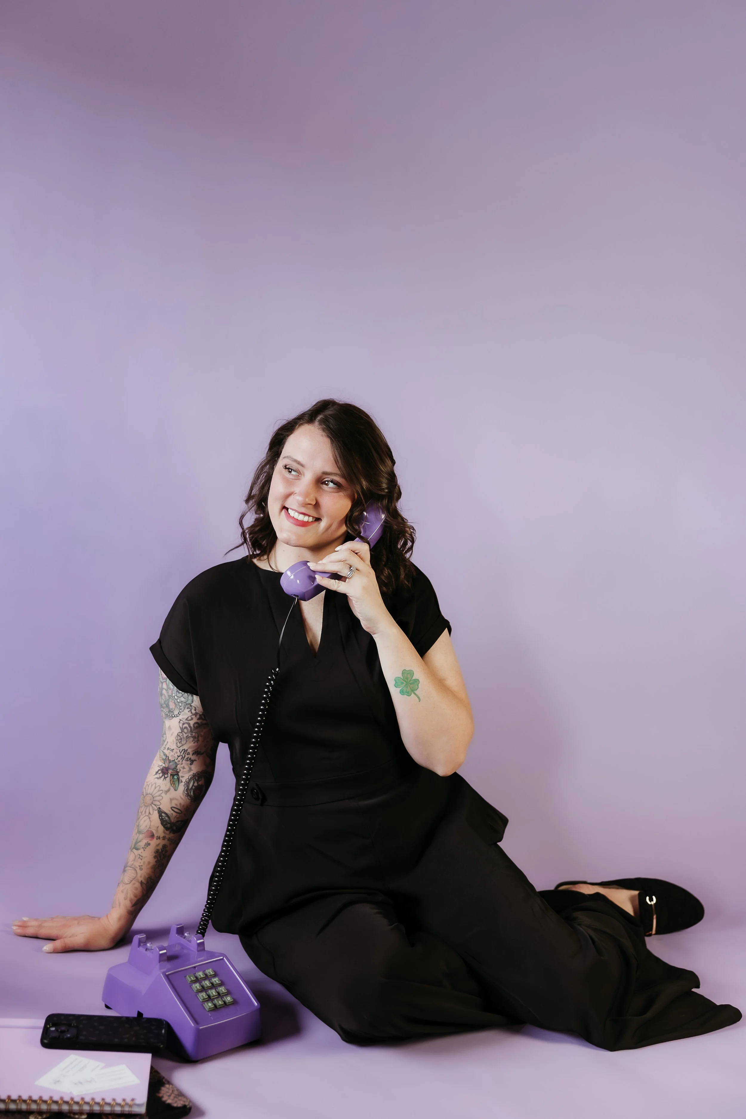 A woman with tattooed arms and dark hair sitting on the floor, holding a purple telephone receiver to her ear, smiling and looking upward, with a purple wall background. There is a purple rotary phone, a cell phone, a notebook, and some papers on the floor.