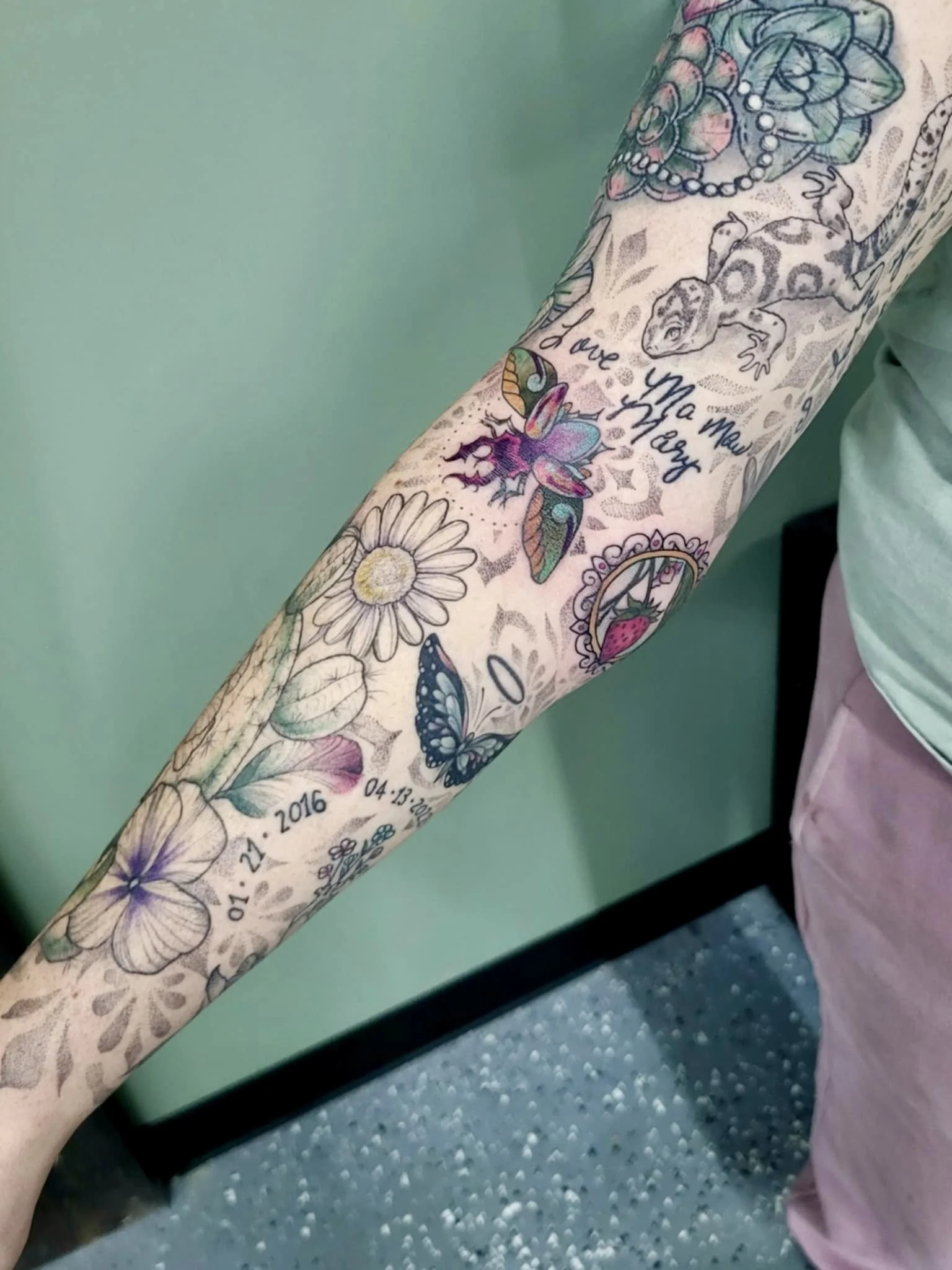 A person's arm with a colorful tattoo sleeve featuring flowers, butterflies, a lizard, and text. The tattoo includes a daisy, a butterfly, a lizard with the text 'Love Ya More,' and dates 01-21-2016 and 04-13-2017.