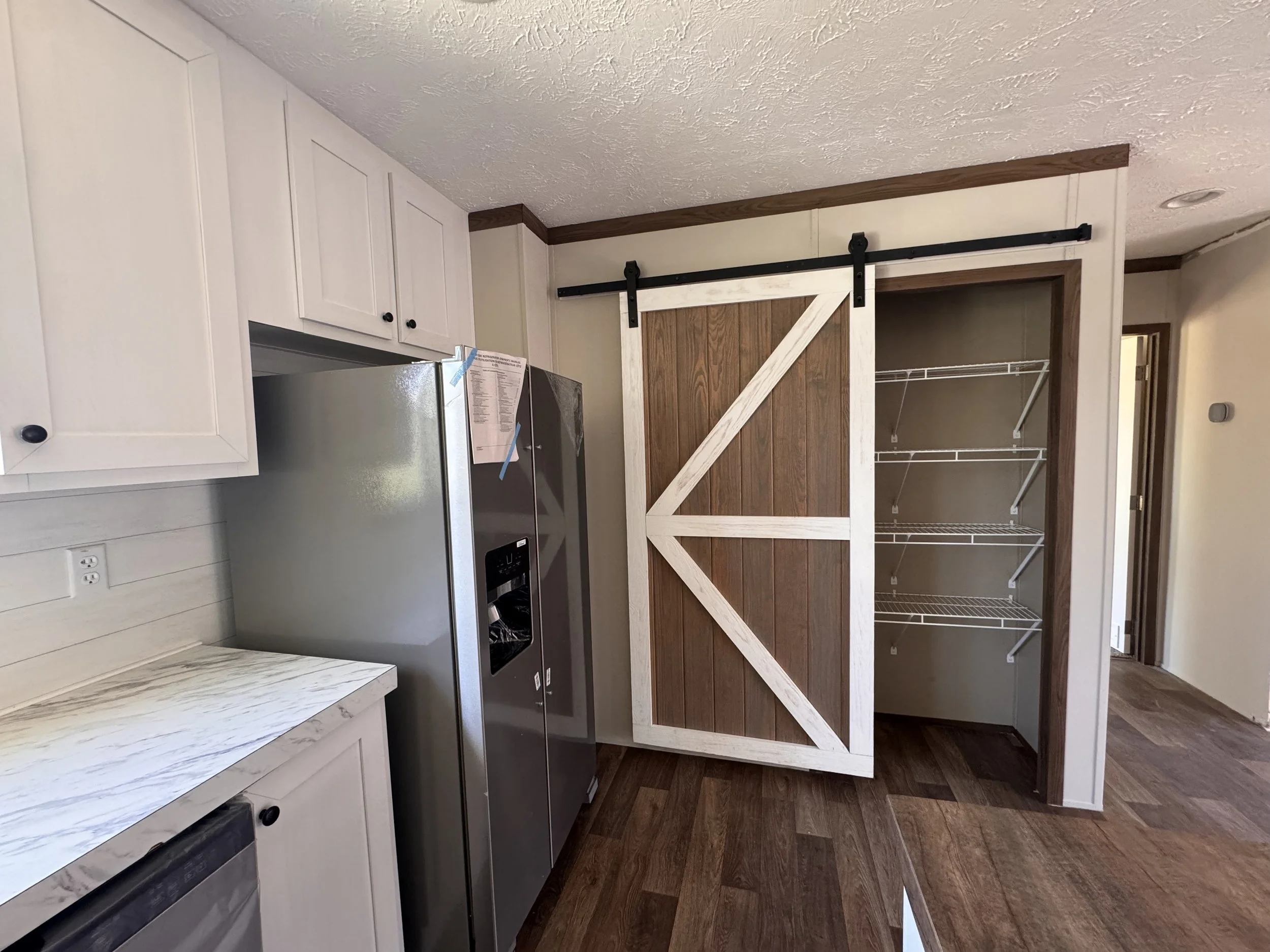 Interior of a room featuring a stainless steel refrigerator, white cabinetry, a rustic sliding barn door on a black track, and a walk-in closet with wire shelving. Hardwood flooring and neutral walls.