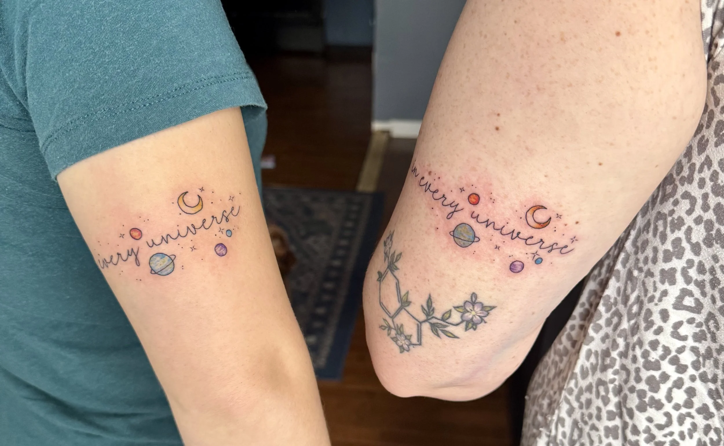 Two people show off matching tattoos on their upper arms featuring planets, stars, and moons with the phrase "in every universe" written in cursive.
