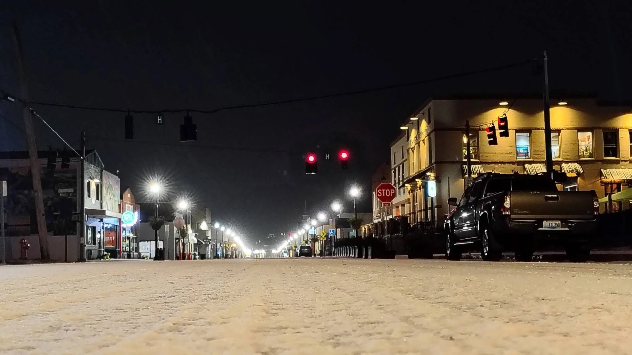 Empty downtown street at night with buildings, streetlights, and parked cars, viewed from ground level.