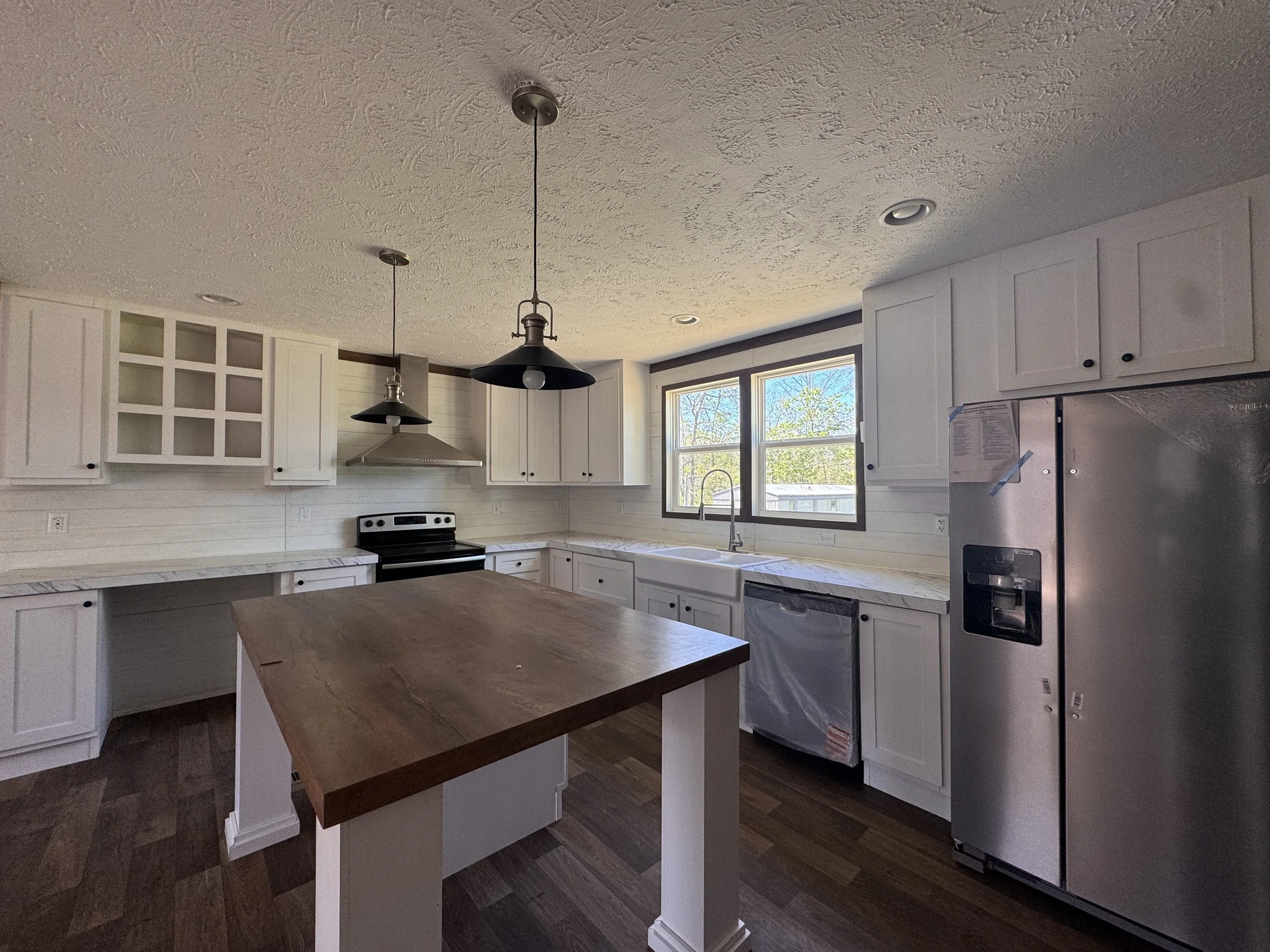 Kitchen with white cabinets, dark wood flooring, a wooden island, and a window letting in natural light. Pendant lighting hangs from the ceiling, and a stainless steel refrigerator is on the right.