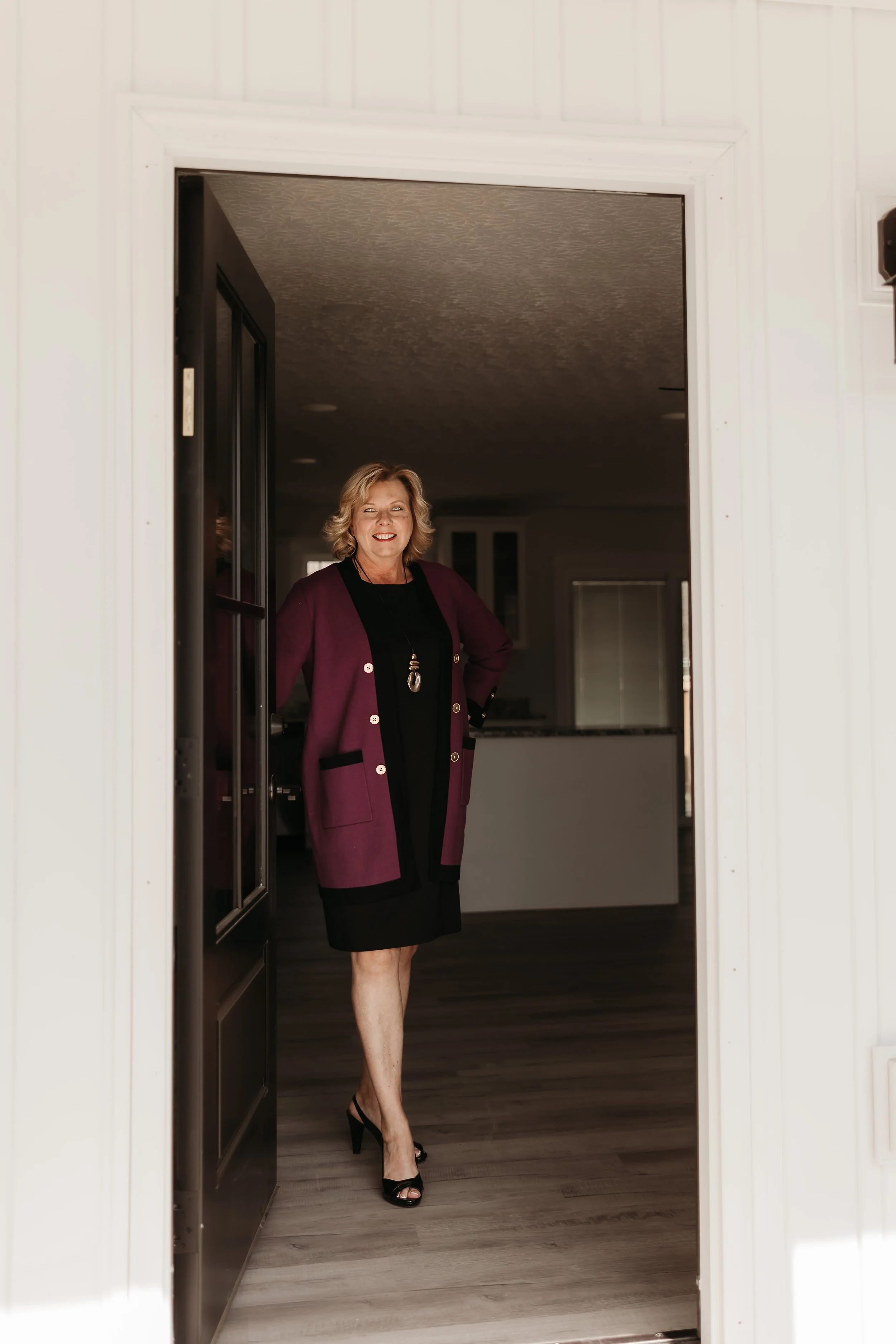 A woman in a black dress and a maroon cardigan standing in a doorway, smiling.