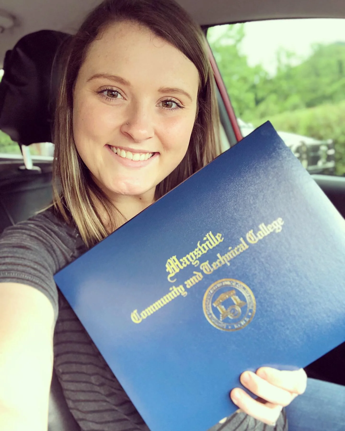 A young woman with brown hair smiling and holding a blue diploma cover with gold lettering that reads 'Mangumite Community and Technical College', sitting inside a vehicle.