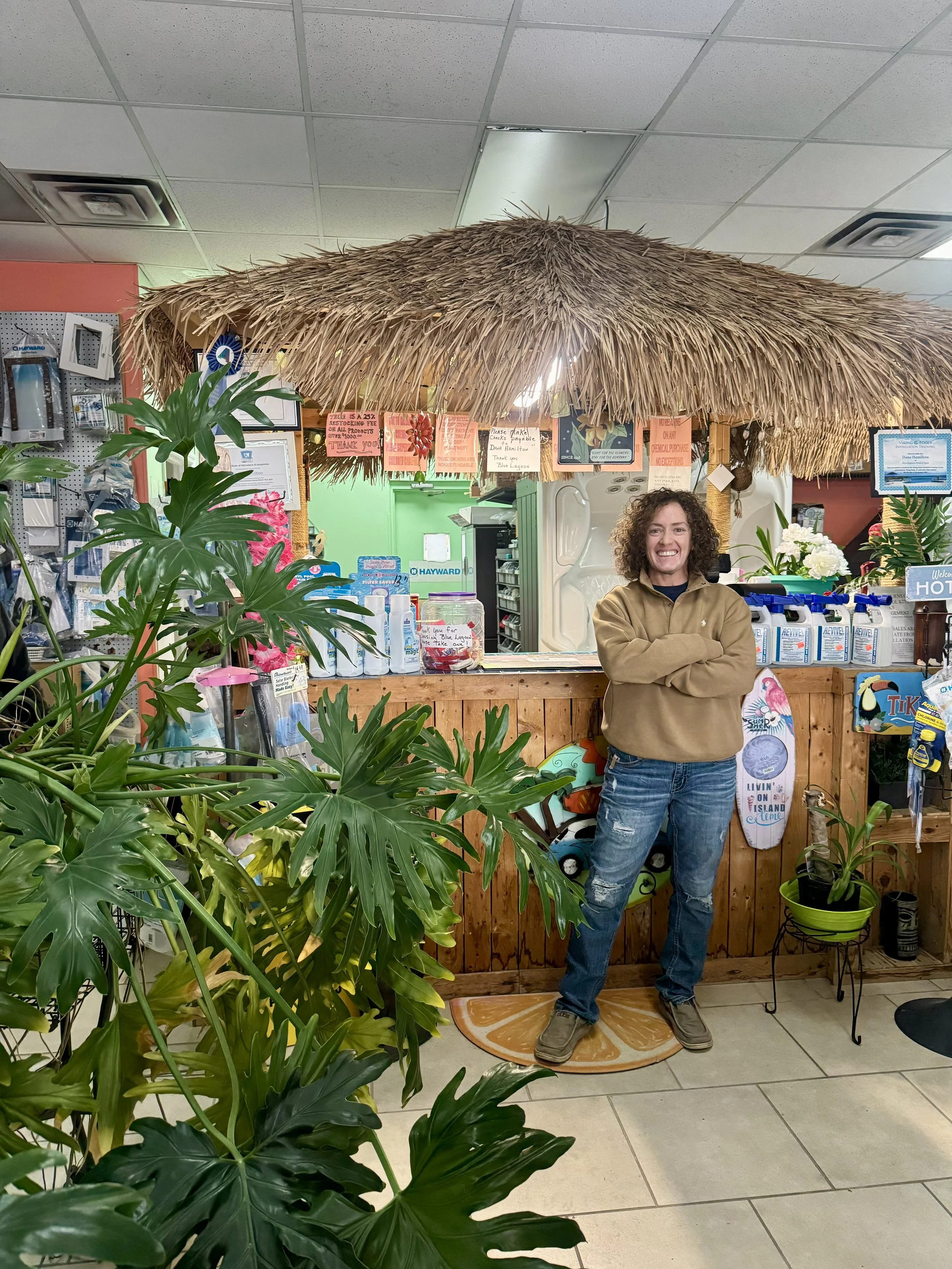 A woman with curly hair standing inside a retail store, smiling with arms crossed. She is standing on an orange and white decorative mat, in front of a wooden counter with a thatched roof overhead. There are green plants around her, and various produ