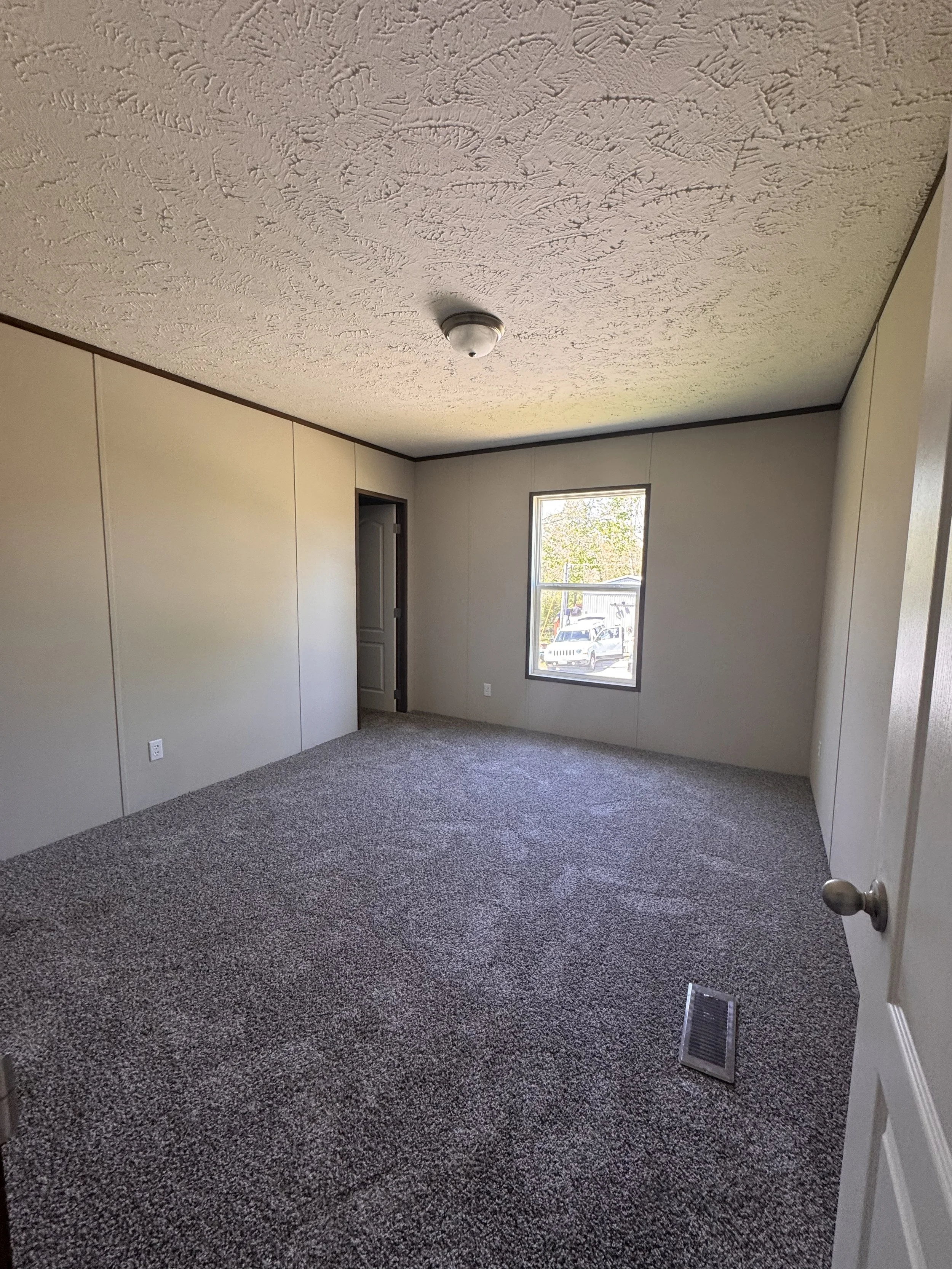 Empty room with beige walls, dark trim, a window showing a parking lot and trees outside, carpeted floor, ceiling with textured pattern, ceiling light fixture, and a vent on the floor.