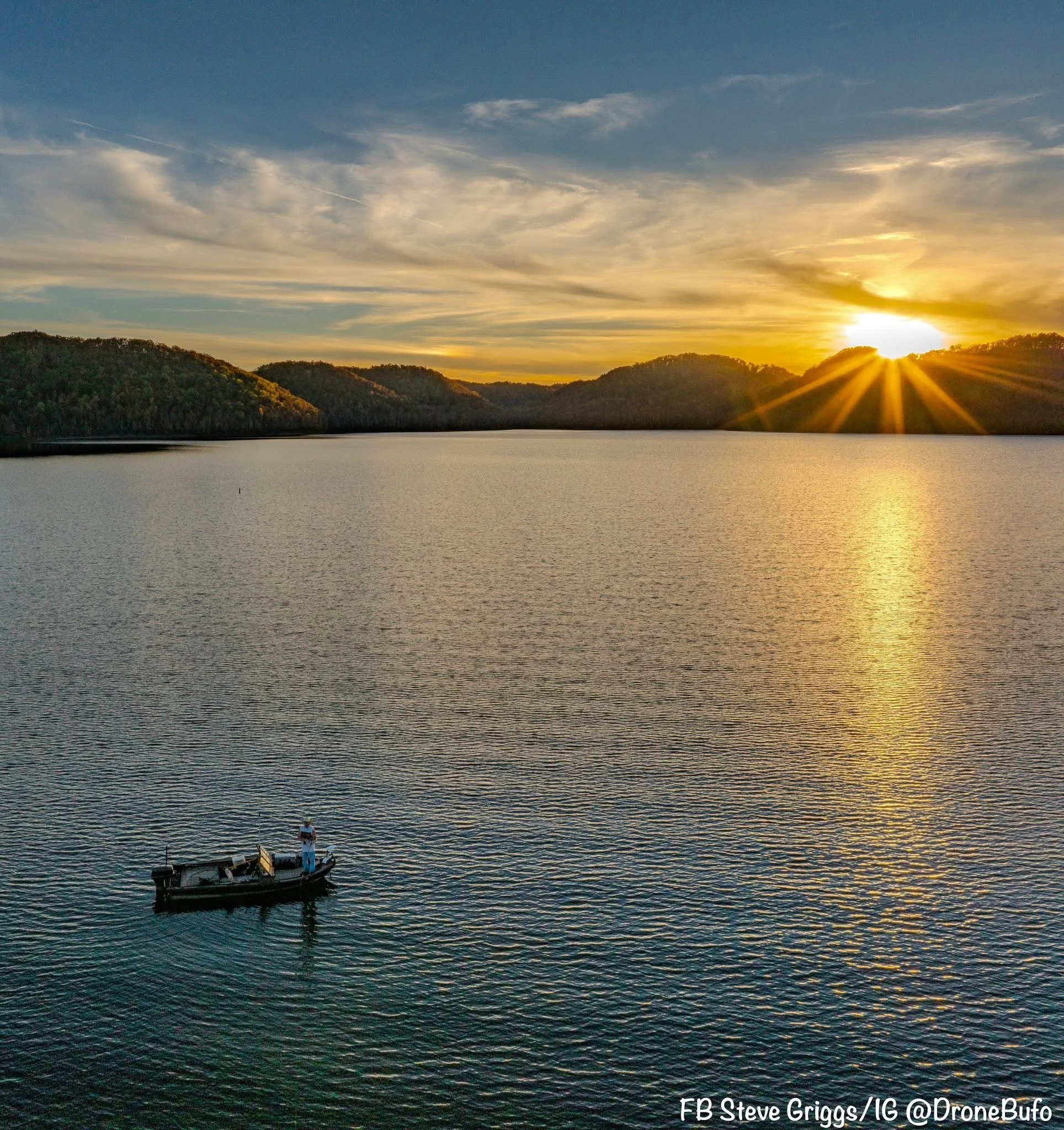 A lake during sunset with a small boat carrying two people in the foreground. The background features hills and a sky with clouds illuminated by the setting sun.