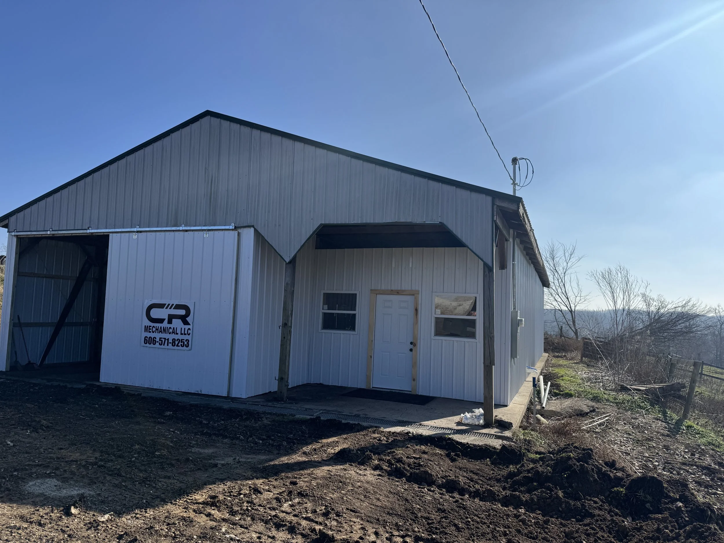 A metal building with a sign that reads 'CR Mechanical LLC' along with a phone number, situated on a dirt lot with some trees and a distant view of hills under a blue sky.