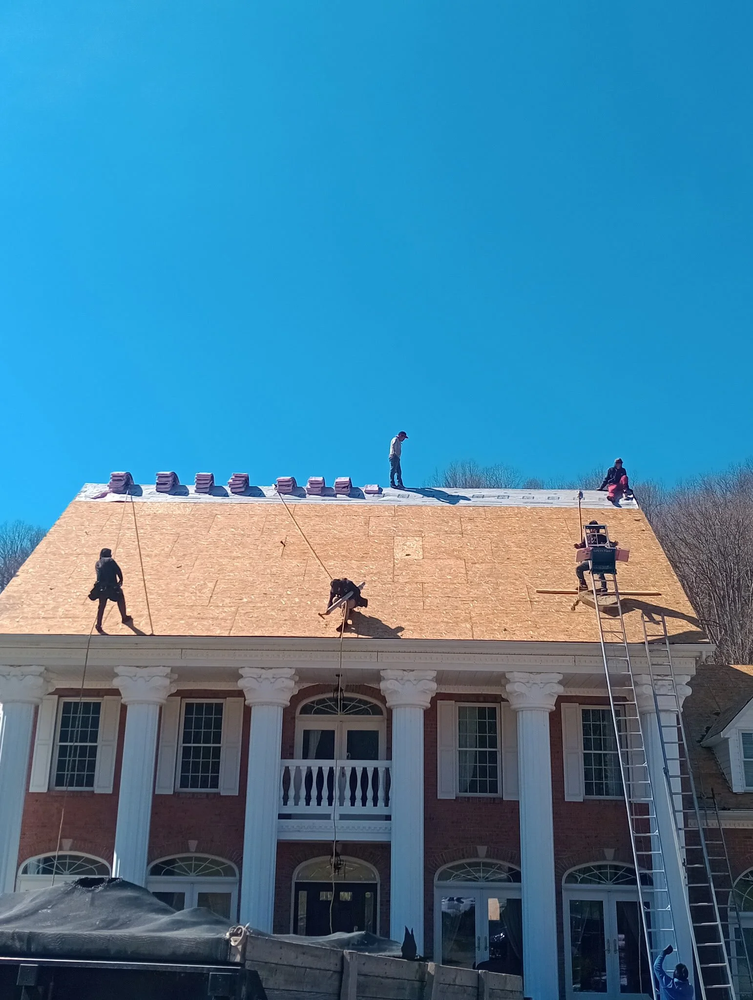 Construction workers installing a new roof on a large brick house with white columns, a balcony, and multiple windows, under a clear blue sky.