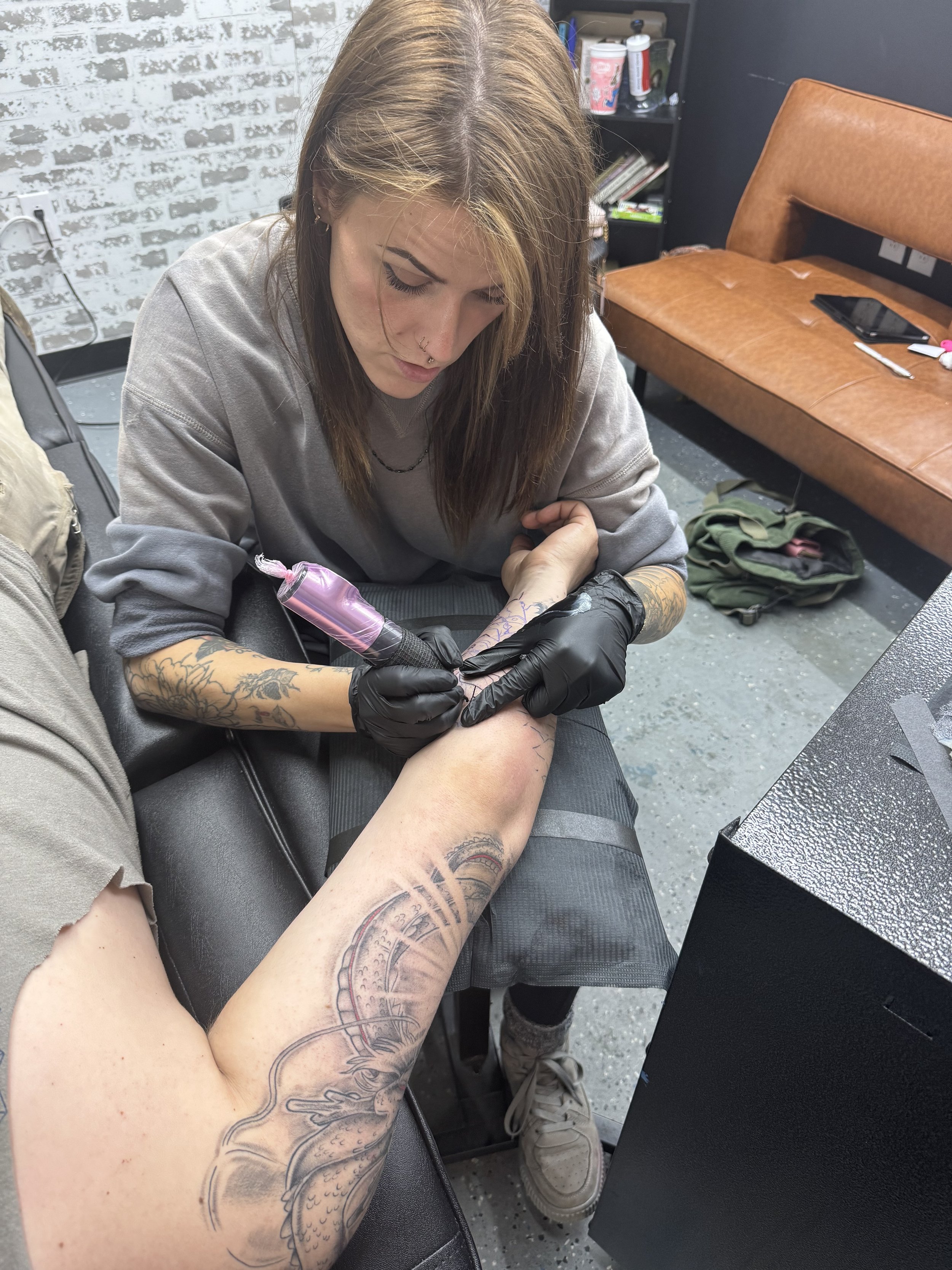 A tattoo artist is making a tattoo on a person's arm. The artist is wearing black gloves and holding a tattoo machine, focusing on her work. The scene is in a tattoo studio with a brick wall background and various supplies on shelves.