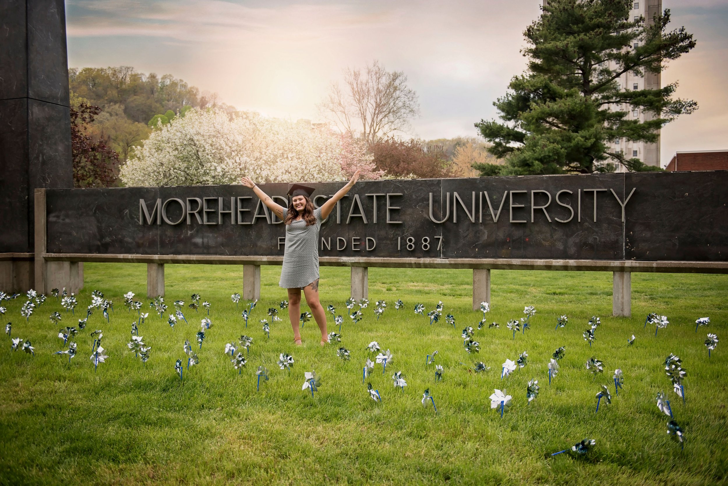 A woman in a graduation gown and cap standing in front of a university sign that reads 'Morehead State University Founded 1887' with her arms raised. She is smiling, surrounded by a green lawn and trees in bloom, during a bright day.