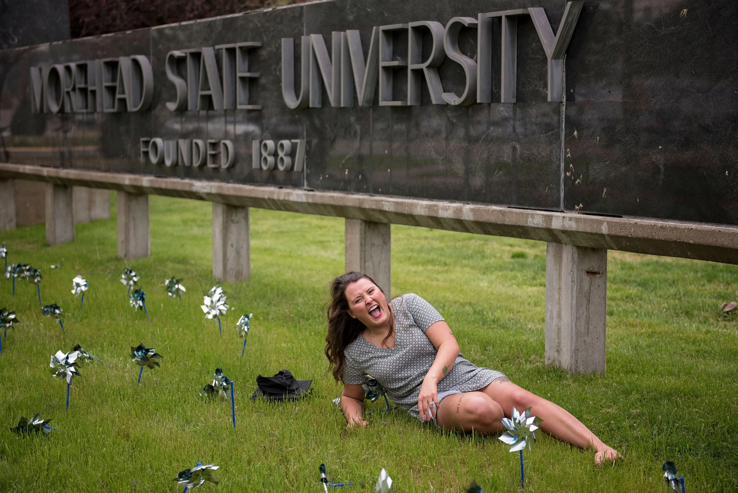 Woman lying on grass, smiling and laughing, near a Memorial of Morehead State University with a large stone sign and pinwheels.