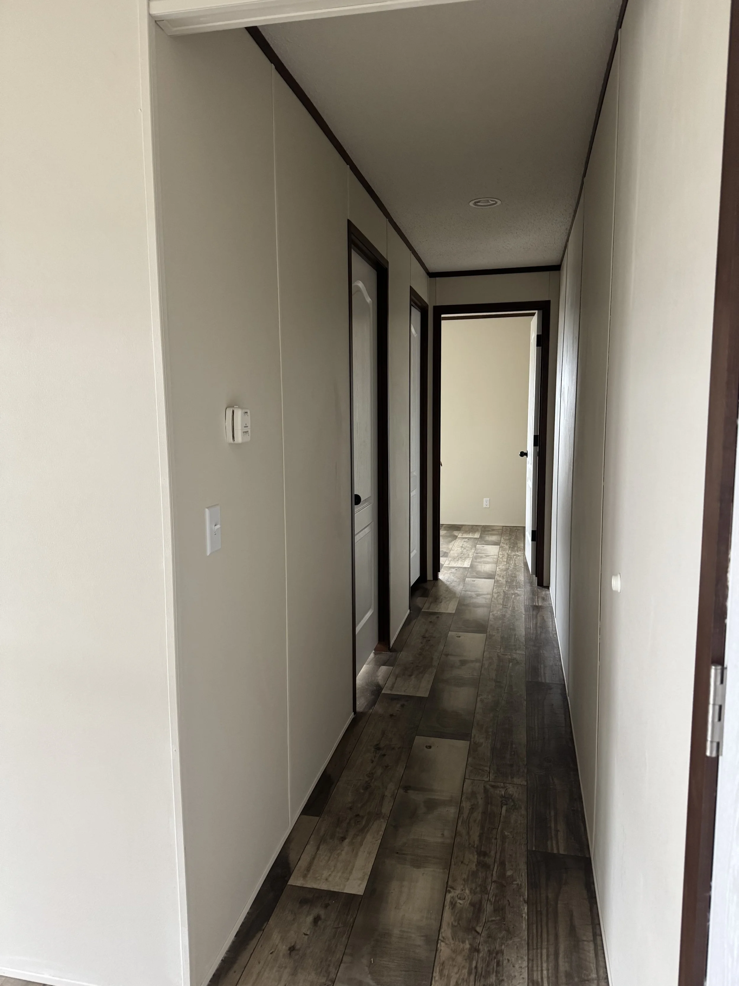 Empty hallway with neutral-colored walls and wood-look flooring, leading to a room at the end.