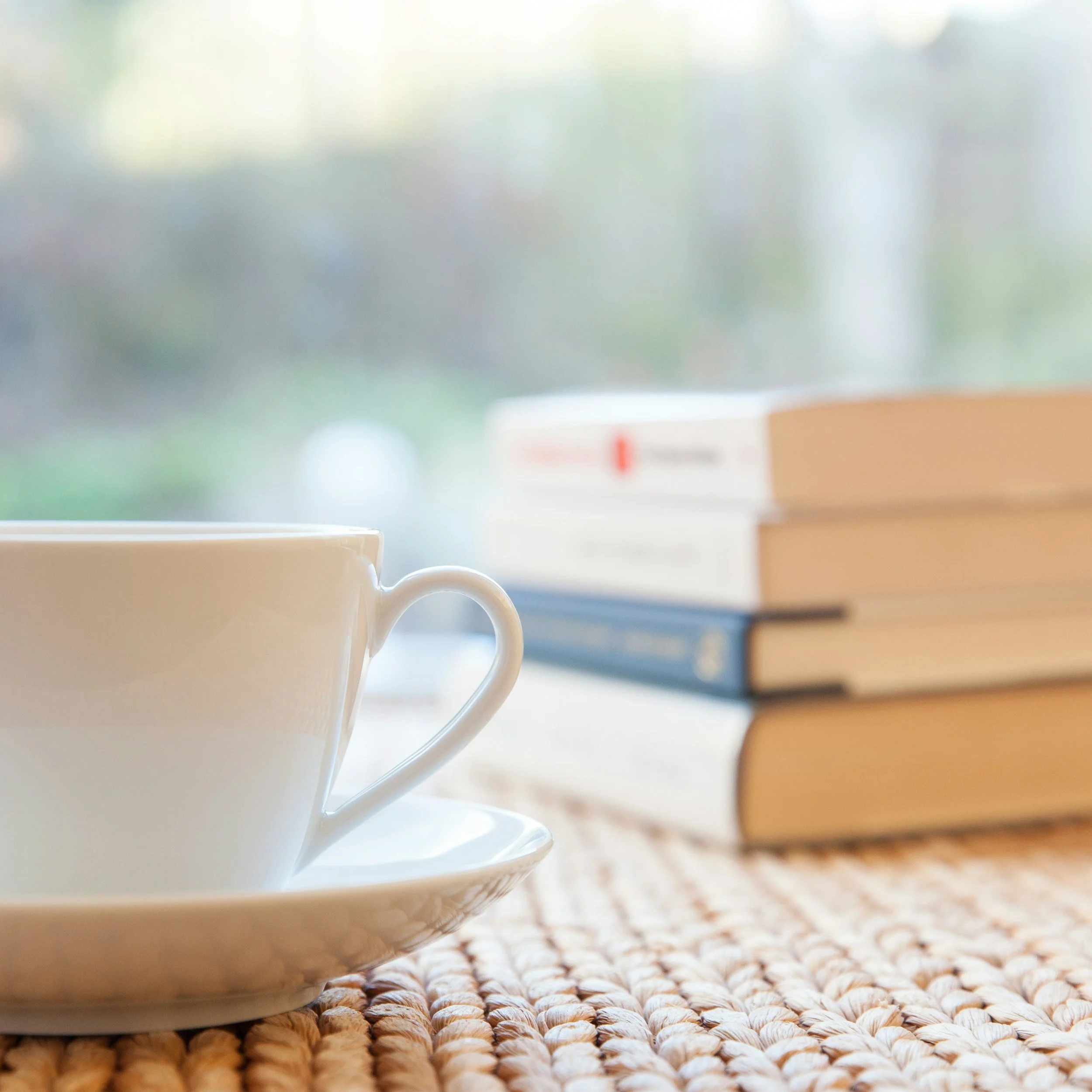 A white coffee cup on a saucer with a stack of books in the background, placed on a woven surface near a window.