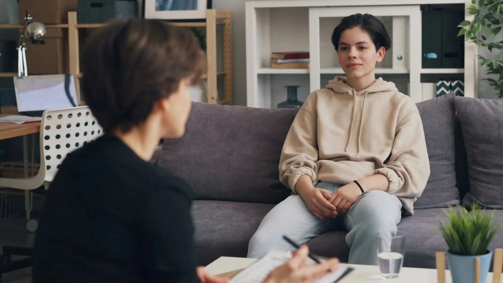 Young woman in a beige hoodie sitting on a gray couch during a therapy session with a therapist taking notes.
