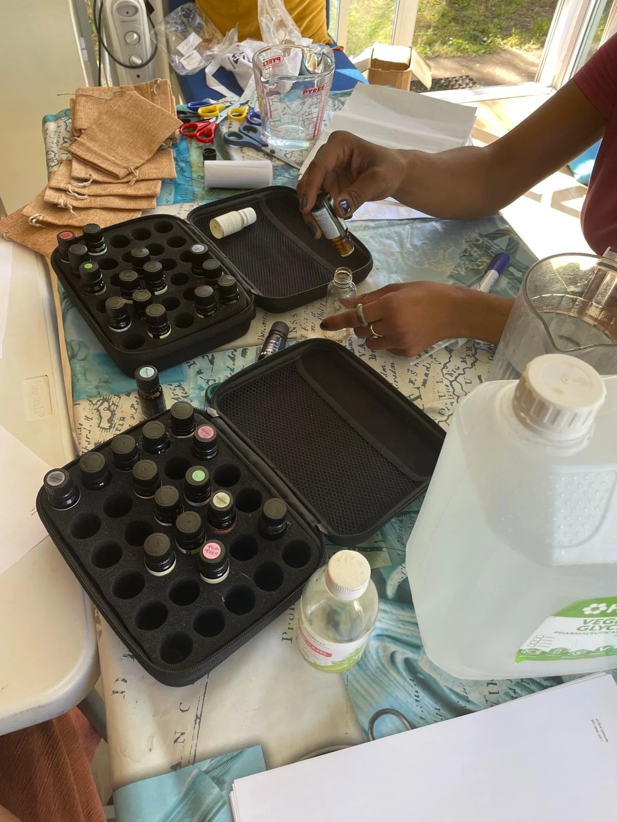 Hands preparing essential oils with small bottles on a table, surrounded by measuring cups, scissors, and cloths.