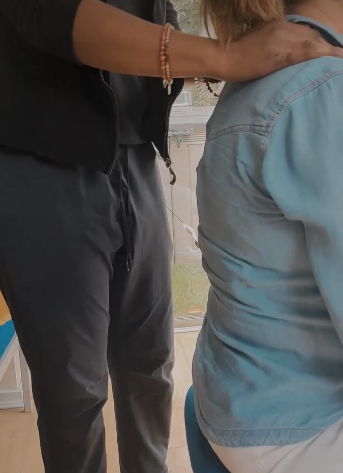 A person in a black jacket and dark pants is standing close to another person sitting down, wearing a light blue denim shirt. The person standing has a beaded bracelet and a wristwatch. The setting appears to be indoors, with a window and some furniture in the background.