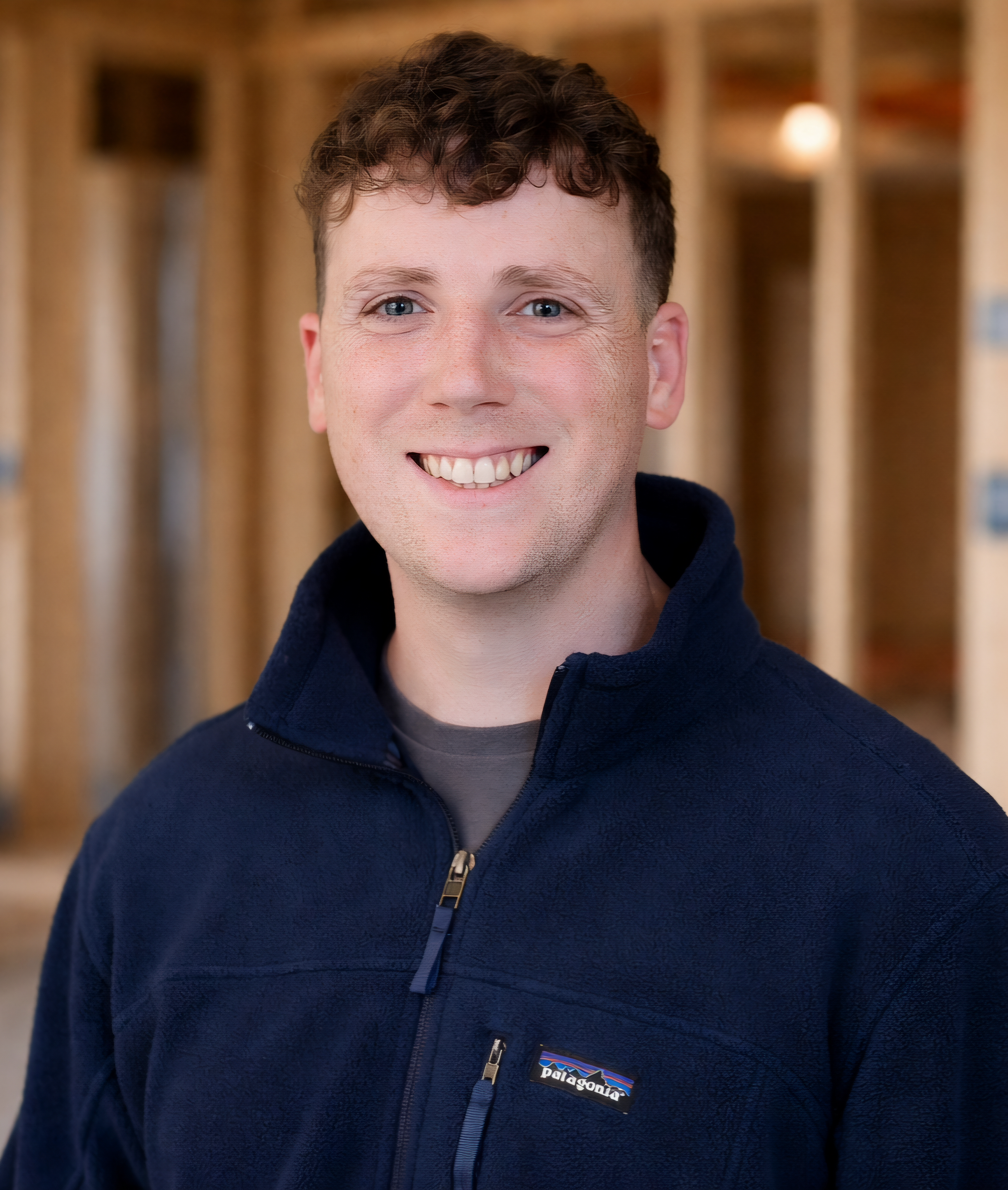 A smiling young man with curly brown hair wearing a navy blue fleece jacket standing in front of a wooden frame background.
