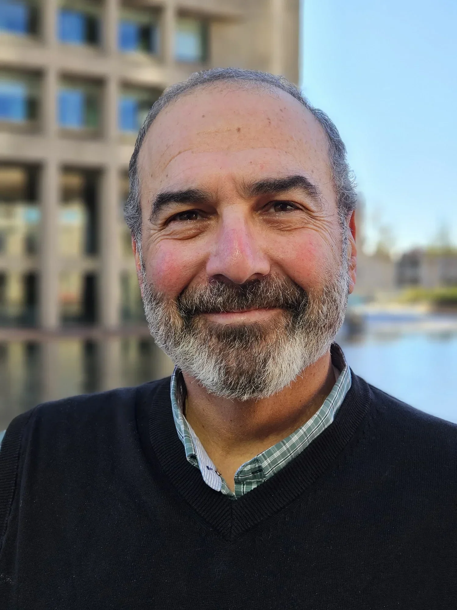 Close-up of a smiling middle-aged man with a beard and short hair, outdoors near water and modern buildings.