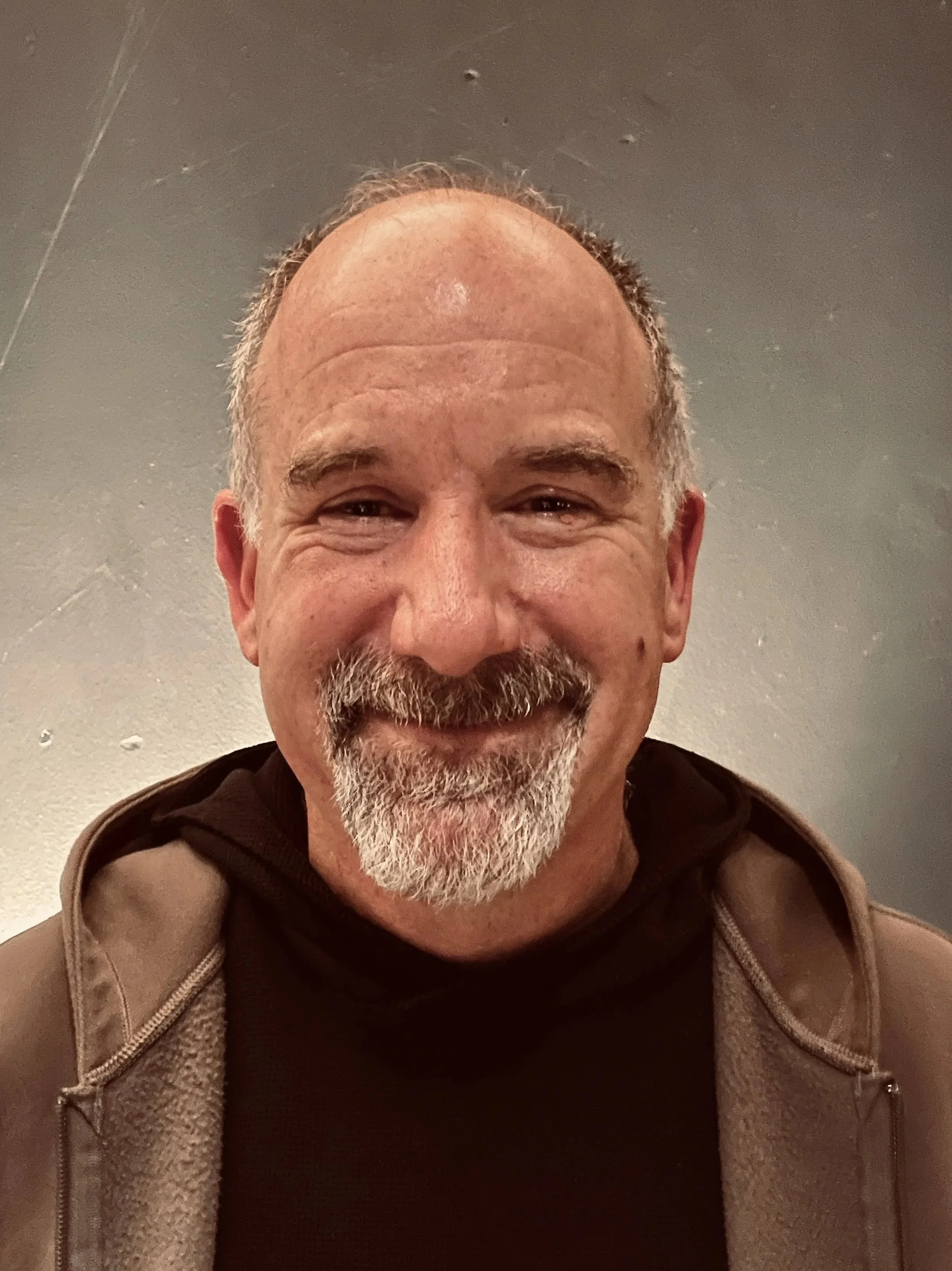 Smiling middle-aged man with a beard and receding hairline wearing a dark hoodie and a light brown jacket, standing against a gray wall.