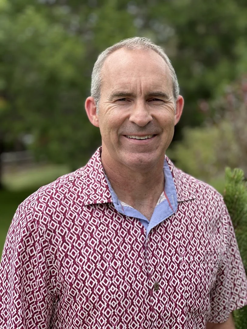 A smiling middle-aged man with short gray hair, wearing a red and white patterned shirt and standing outdoors with greenery in the background.