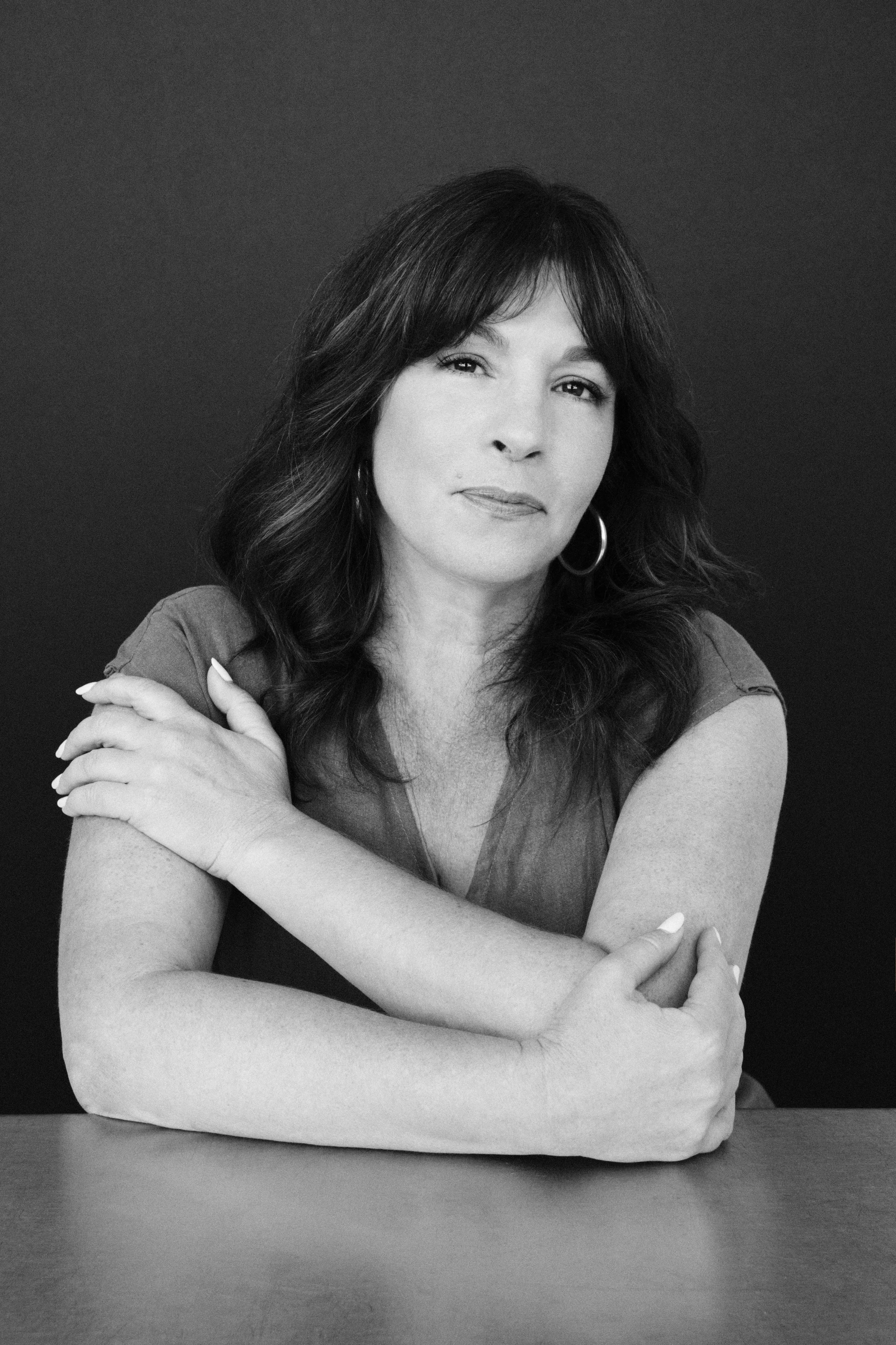 Black and white portrait of a woman with dark wavy hair, wearing hoop earrings, sitting with her arms crossed on a table, against a dark background.