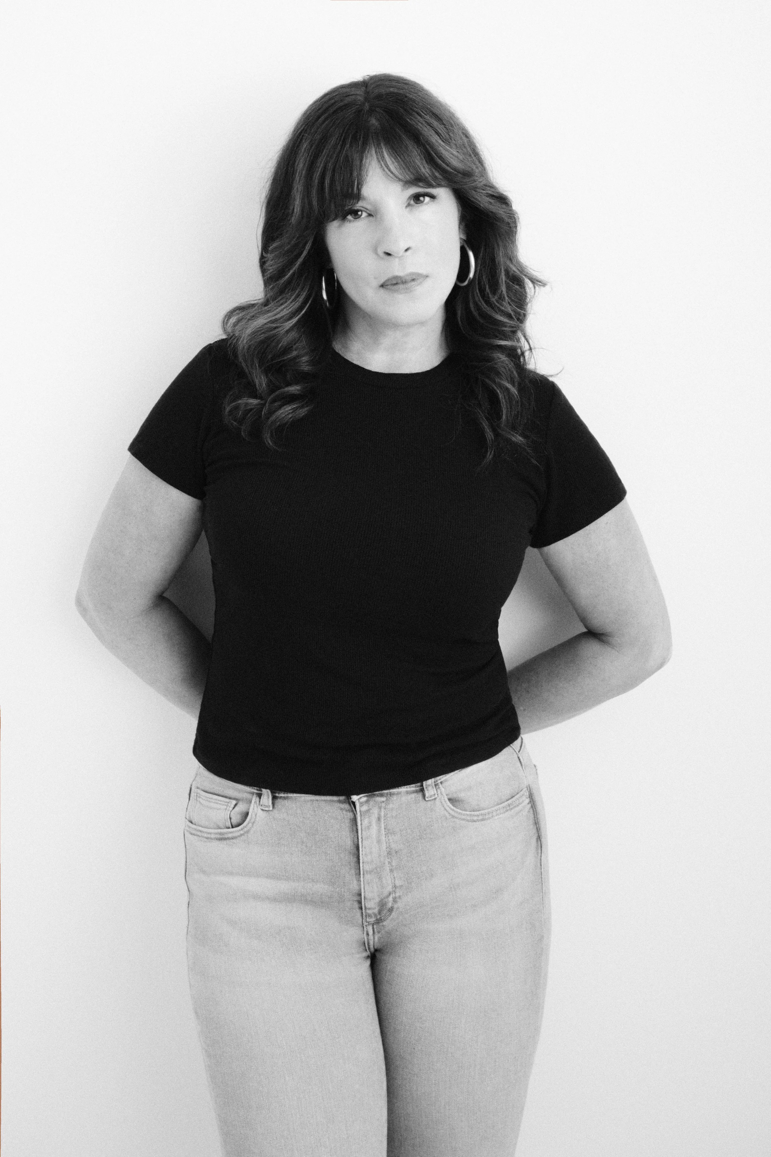 Black and white portrait of a woman with shoulder-length wavy hair, wearing hoop earrings, a black t-shirt, and light-colored jeans, standing against a plain background.