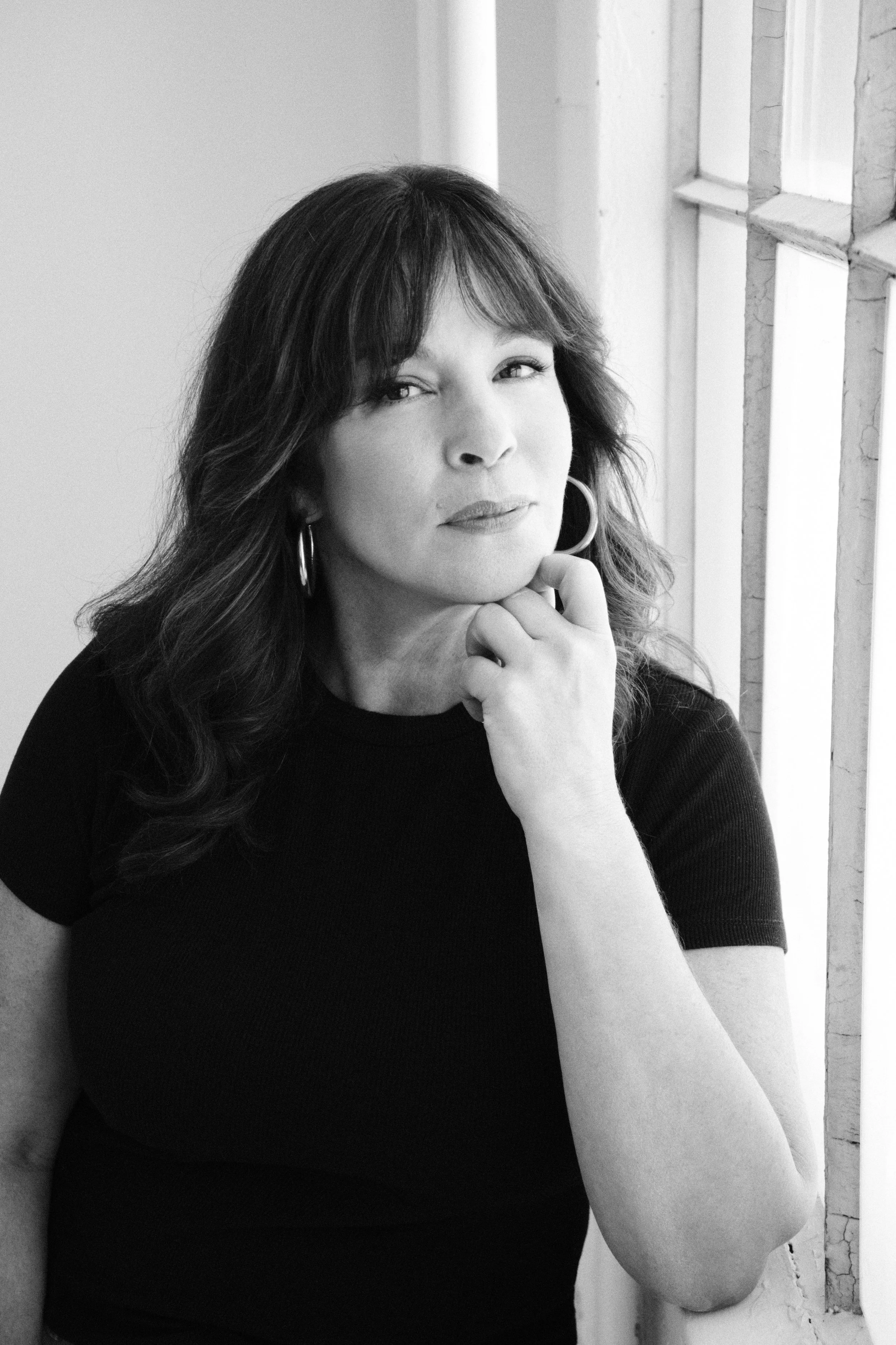 Black and white portrait of a woman with wavy hair, wearing hoop earrings, looking at the camera with one hand near her chin, near a window with peeling paint.