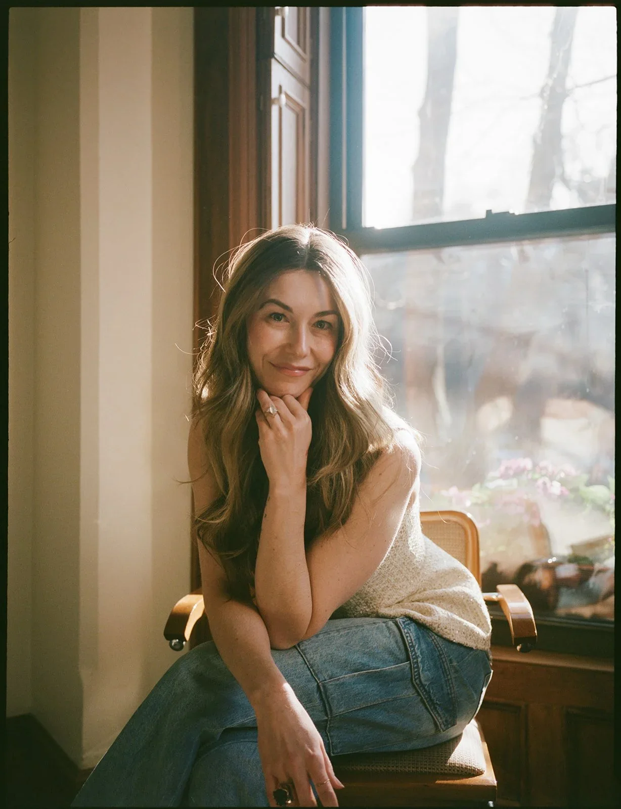 A woman with long, wavy hair sitting on a wooden chair by a window with sunlight streaming in, wearing a sleeveless top and jeans, looking at the camera with a soft smile.