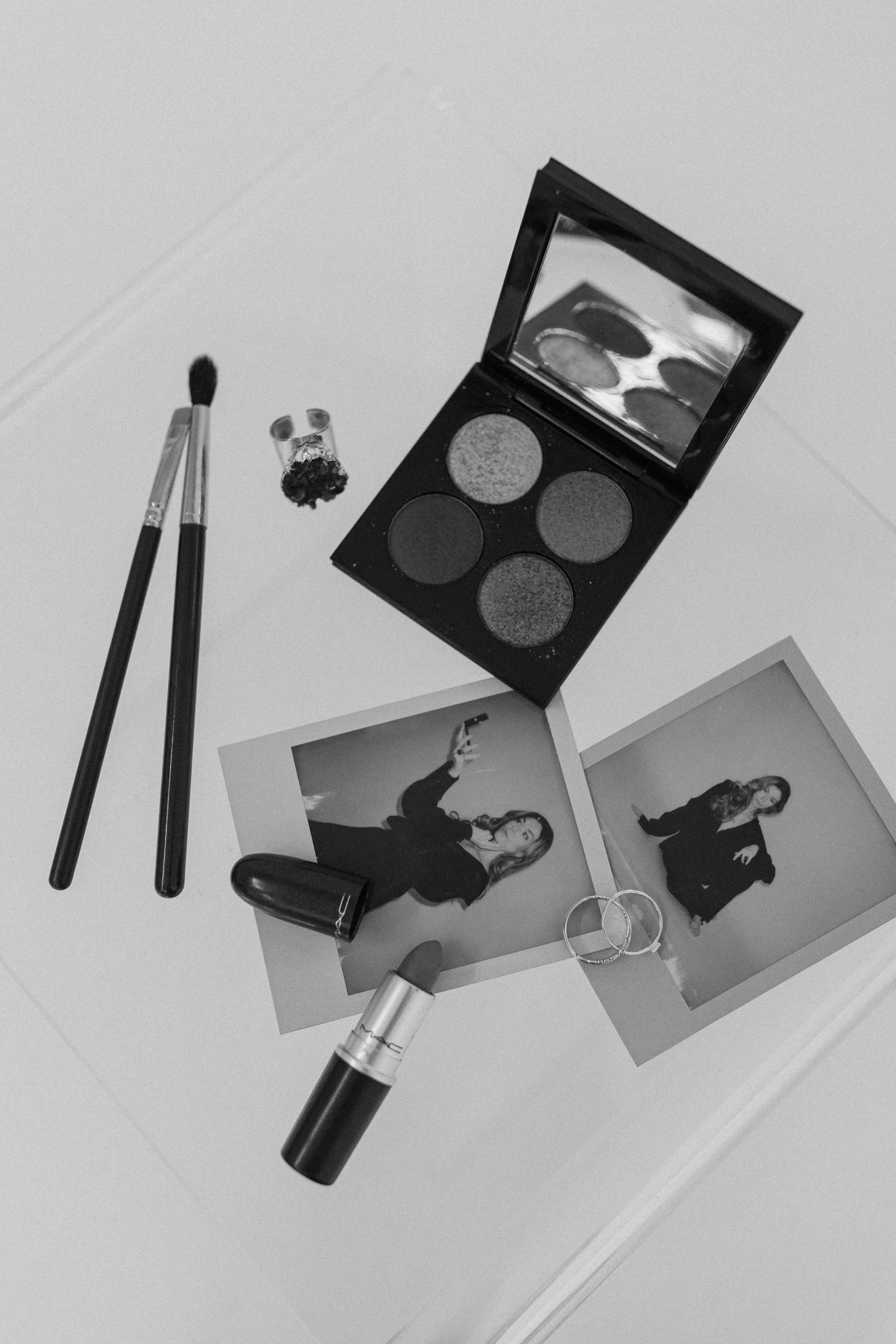 Black and white photo of makeup items, jewelry, two Polaroid photos of a woman, and an open eyeshadow palette on a flat surface.