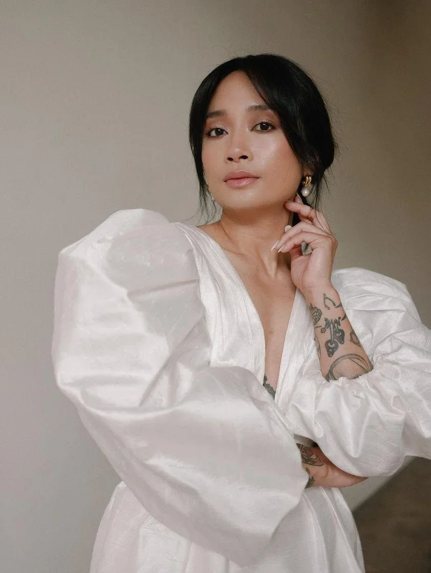 A woman with short black hair and light makeup, wearing pearl earrings and a white puffy-sleeve dress, posing against a plain wall, with her right hand touching her chin and her left arm crossed over her chest, showing tattoos on her forearm.