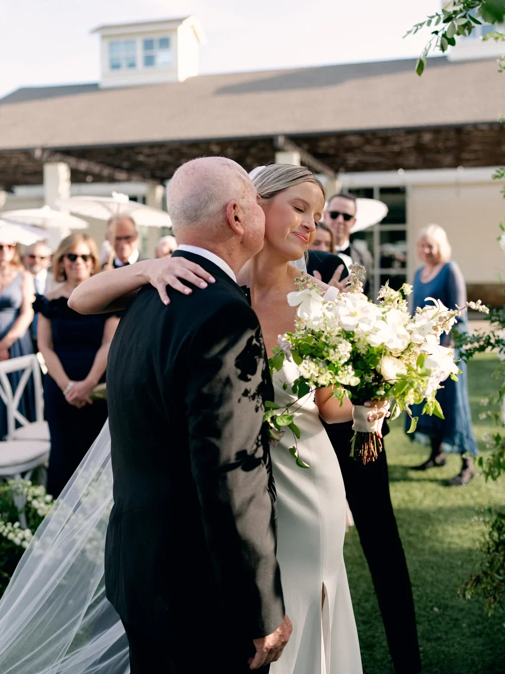 A bride in a white wedding dress holding a bouquet of white flowers is being embraced by an older man in a black tuxedo during an outdoor wedding ceremony. Guests are standing in the background watching and smiling.