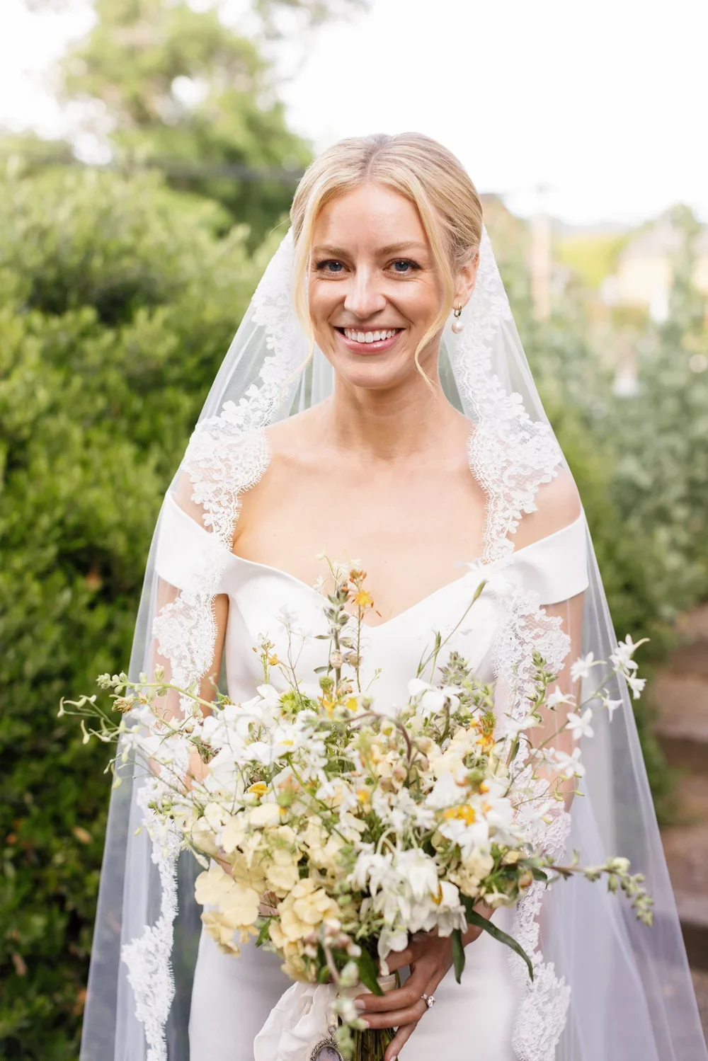 A smiling bride in a white wedding dress with lace details, wearing pearl earrings, and holding a bouquet of white and yellow flowers outdoors on a sunny day.