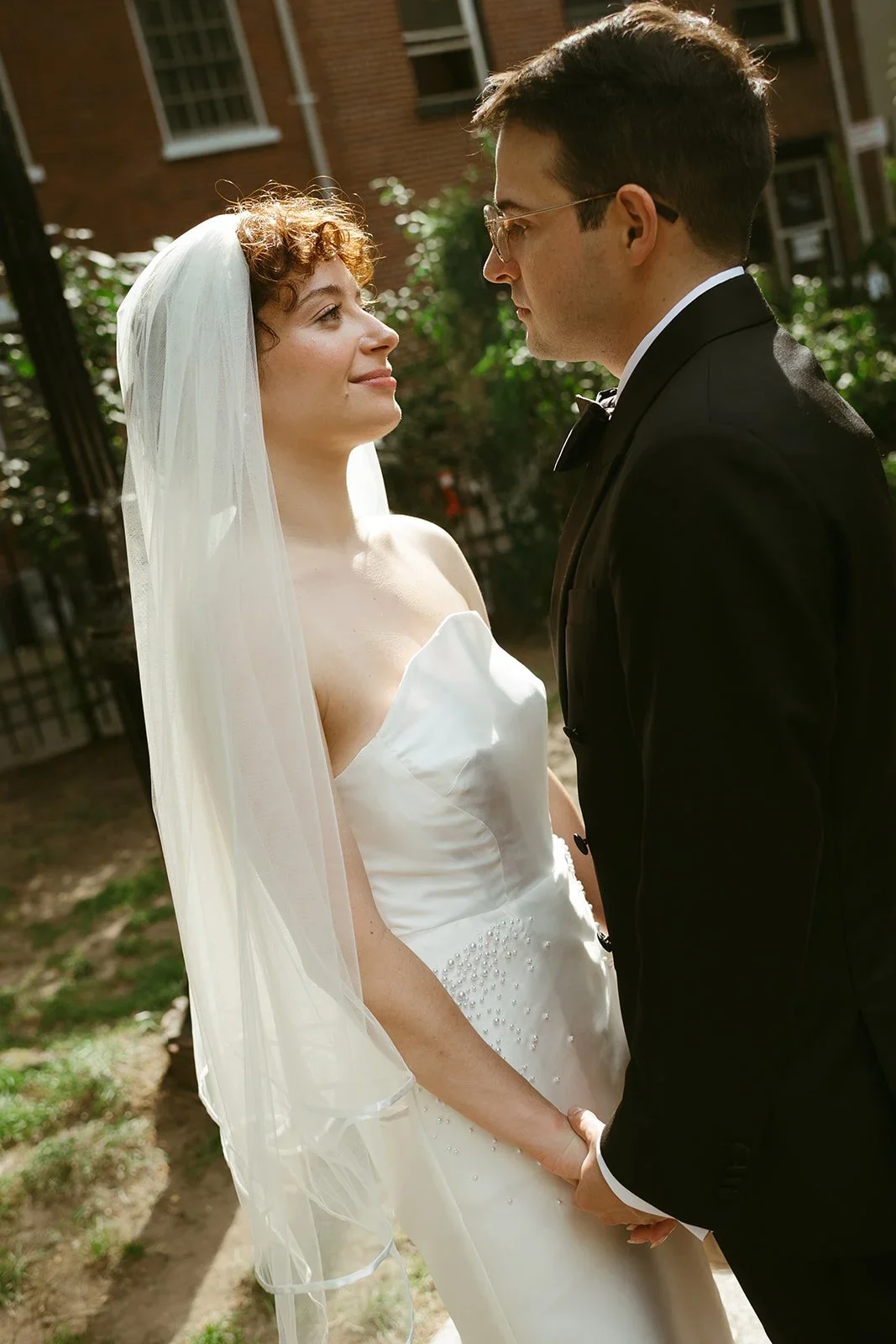 A bride and groom stand face-to-face outdoors, holding hands, with a brick building and greenery in the background.