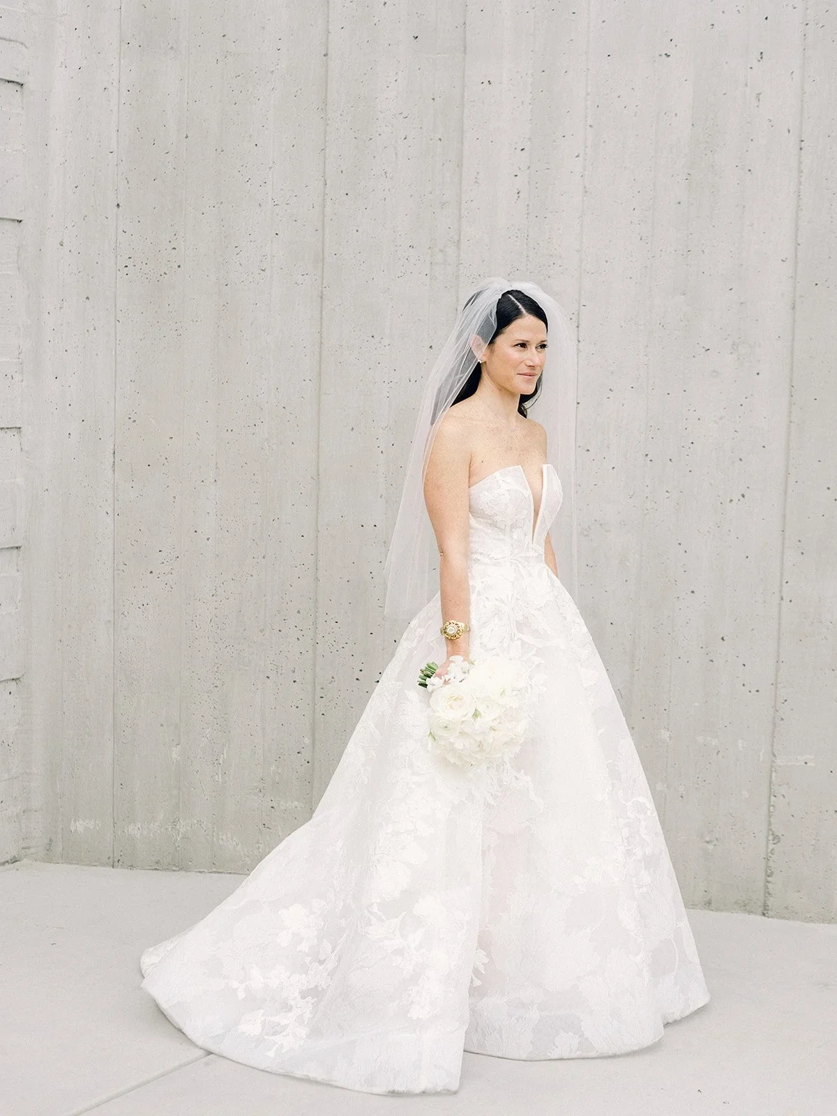 Bride in a strapless white wedding gown holding a bouquet of white flowers, standing against a concrete wall with a veil on her head.