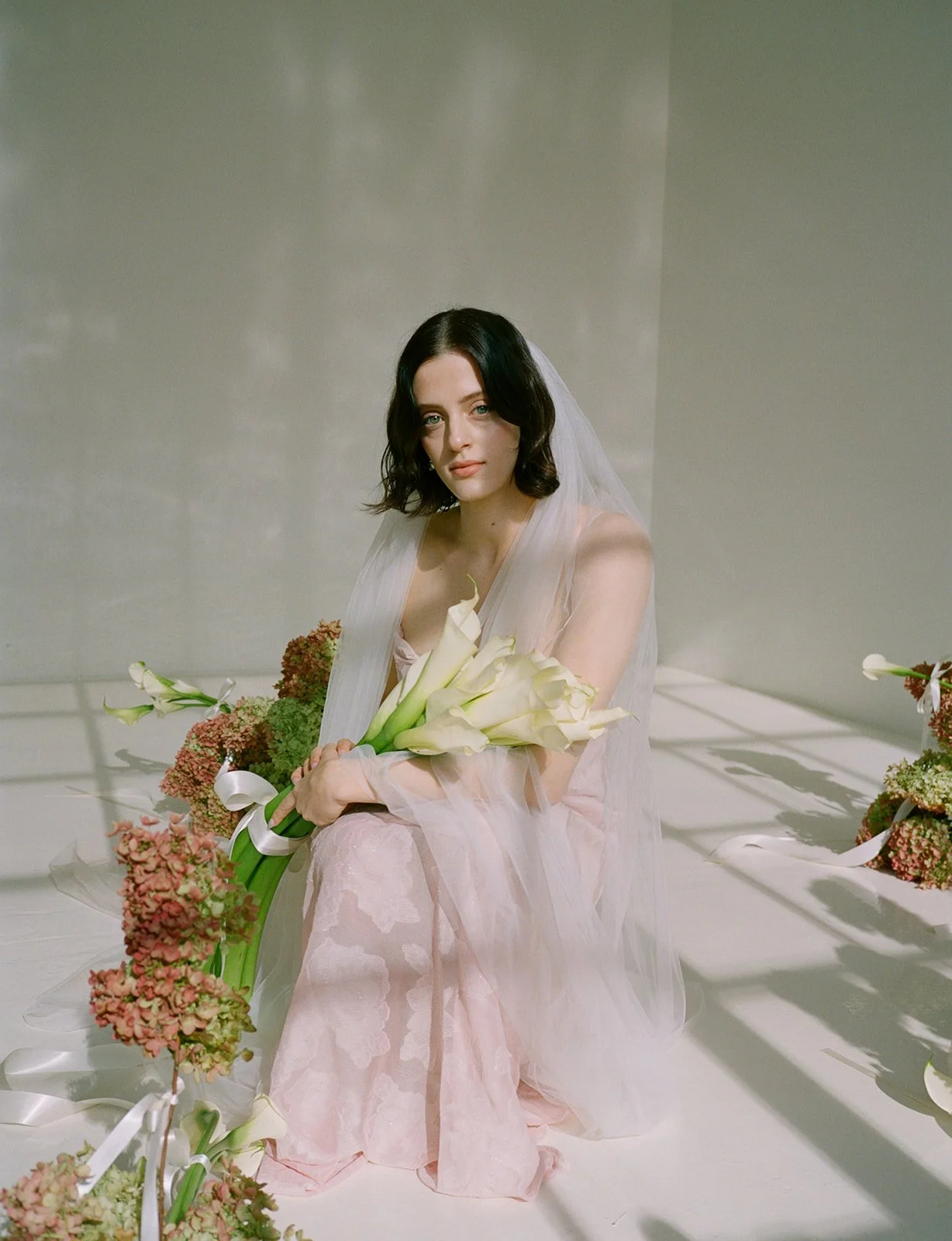 A woman with shoulder-length dark hair in a soft pink and sheer dress, sitting on a white surface, holding a bouquet of white calla lilies, surrounded by floral arrangements and ribbons, with shadows cast on the backdrop.