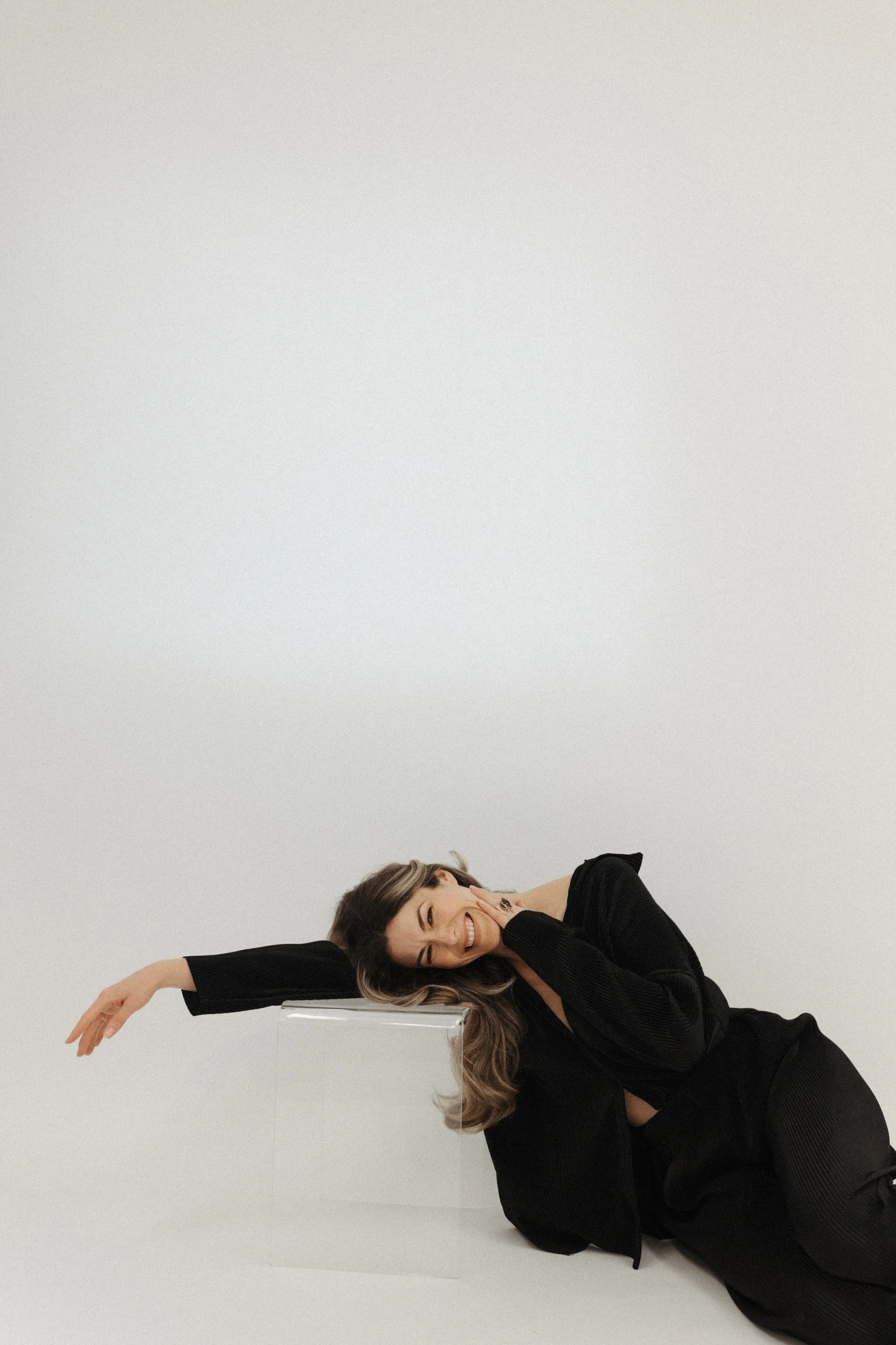 Woman wearing black outfit smiling and lying on the floor with her head resting on a clear rectangular block, in a minimalistic white studio setting.