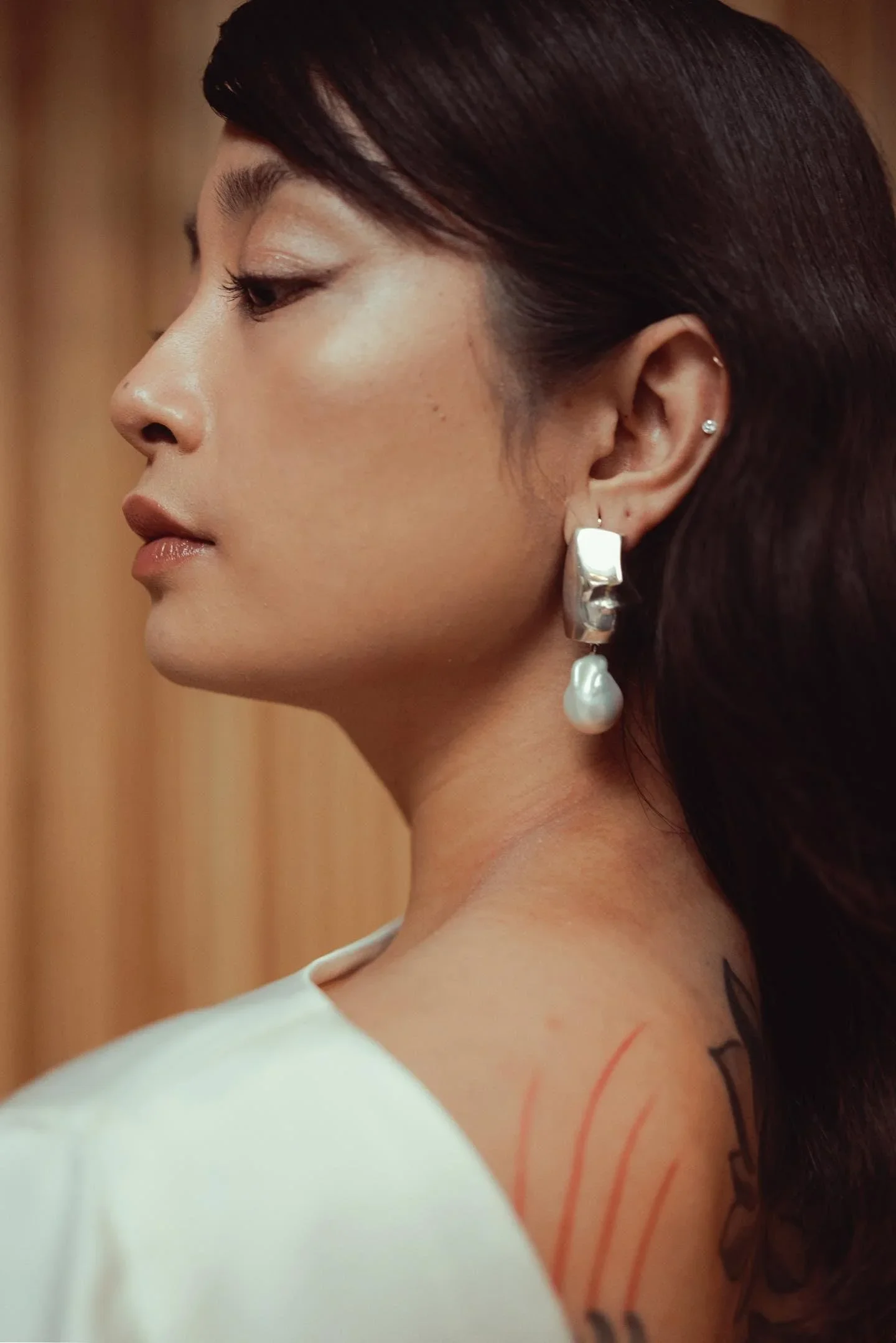 Close-up side view of a woman with dark hair, wearing earrings with a pearl drop, and a tattoo on her shoulder. She appears to be in a softly lit indoor setting.