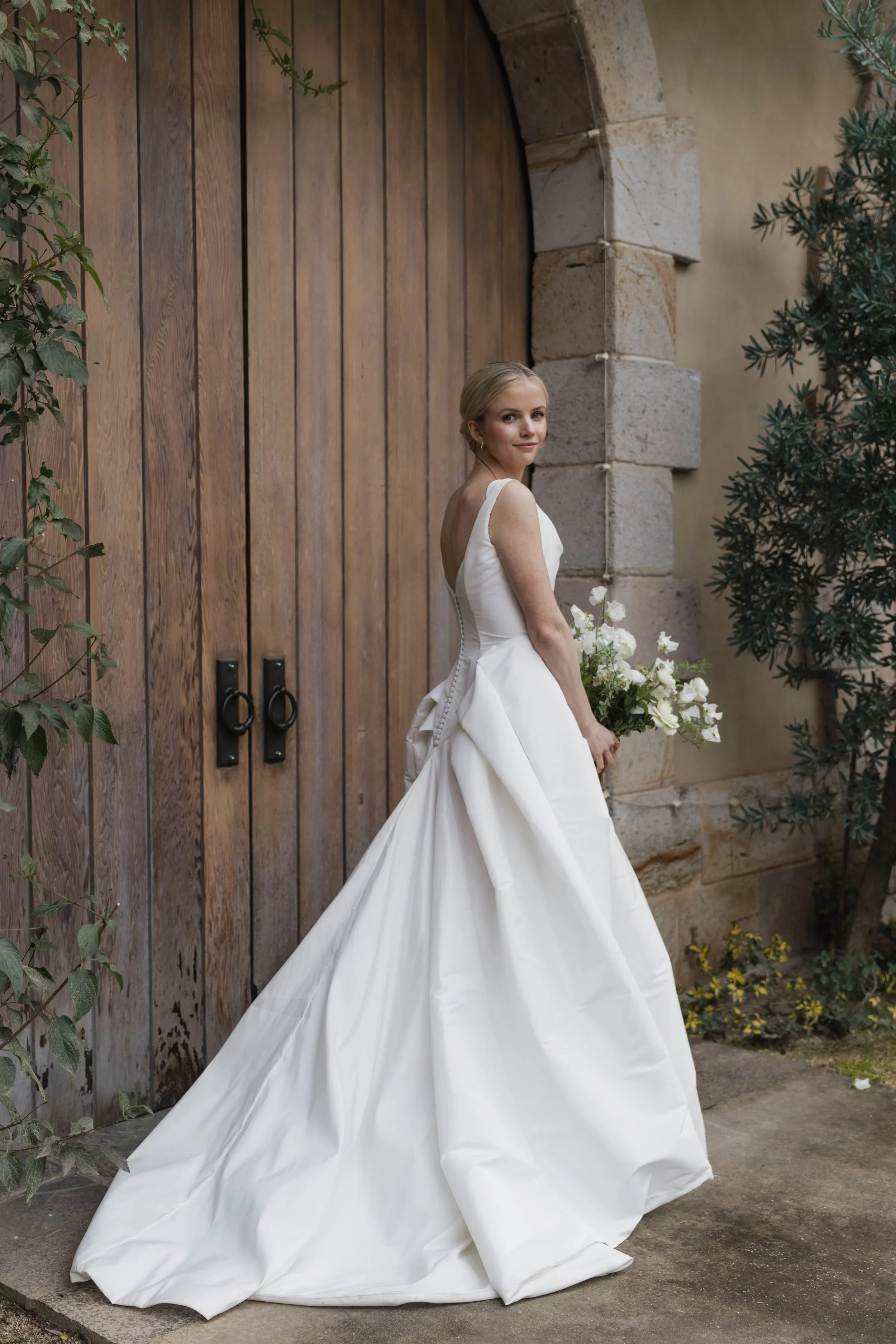 A bride in a white wedding dress holding a bouquet of white flowers, standing outside near a wooden door and stone wall, smiling at the camera.