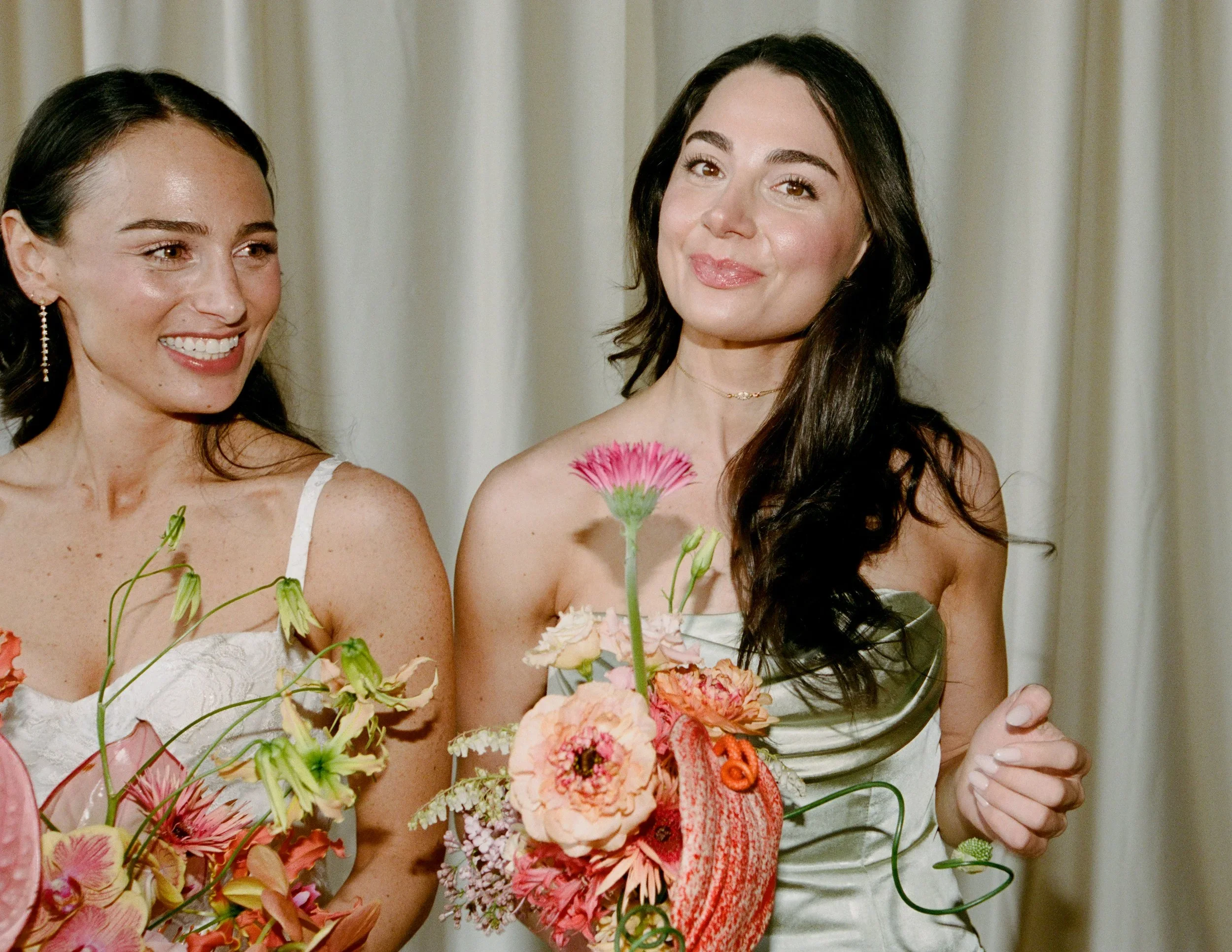Two women in white dresses holding bouquets of pink and peach flowers, standing in front of a cream-colored curtain, smiling.