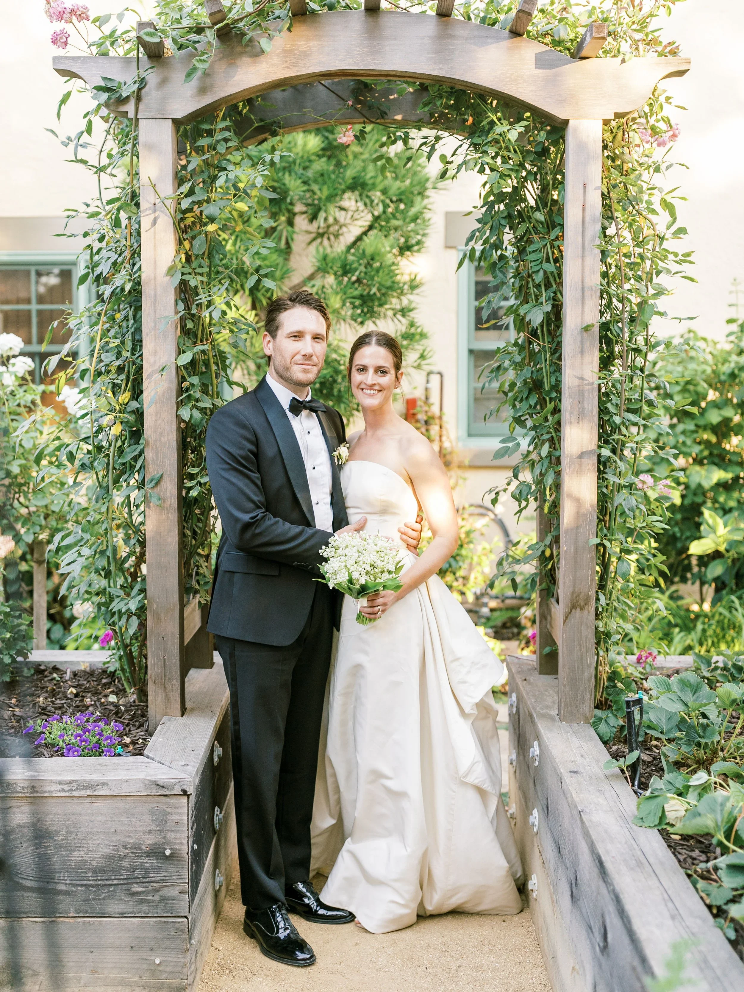 A bride and groom standing together under a wooden arch at their wedding, with flowers and greenery around them.