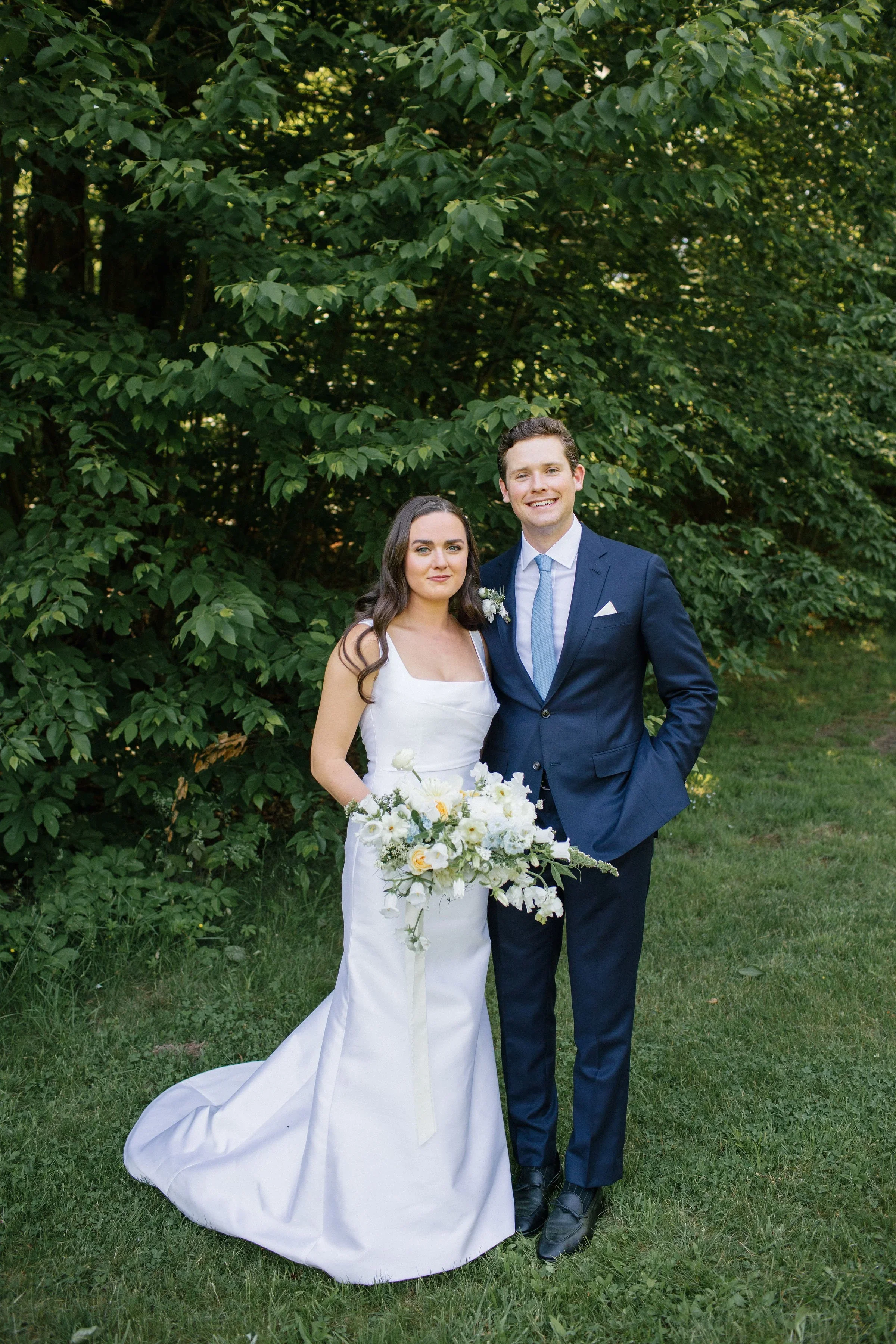 Bride and groom standing outdoors on grass in front of lush green foliage, with bride in white wedding gown holding a large bouquet, and groom in a dark blue suit with light blue tie, smiling at the camera.