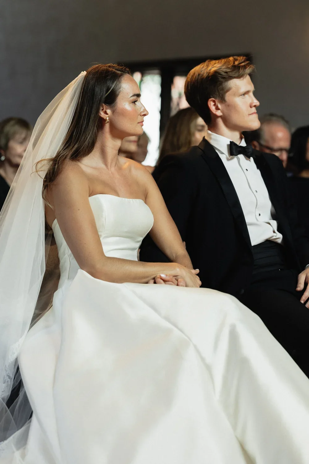 Bride and groom sitting side by side during a wedding ceremony, with the bride wearing a strapless white wedding dress and veil, and the groom in a black tuxedo with a bow tie.